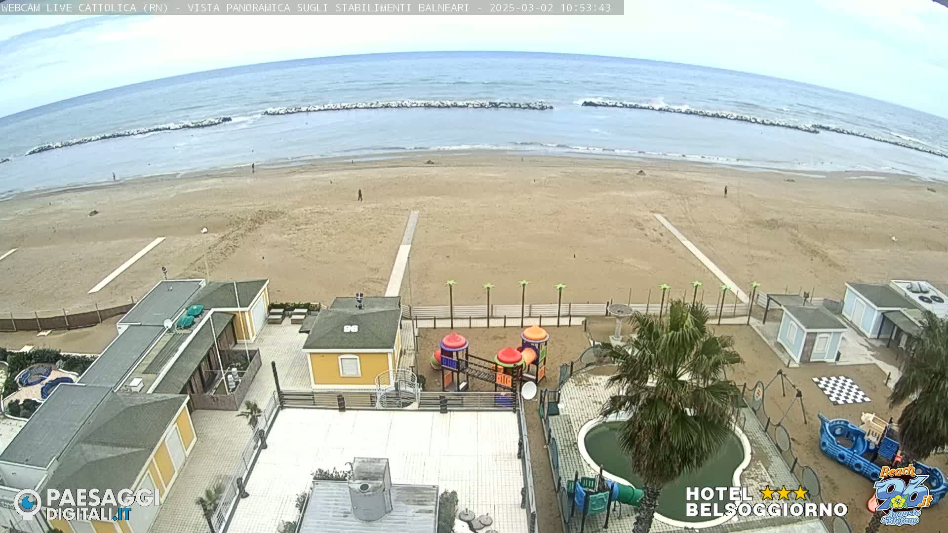 An overcast day at a beach shows a sandy shore, calm ocean waves, beach buildings, and a playground.