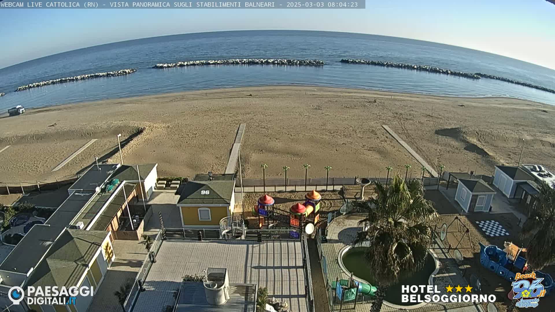 A beach with calm, clear water and a mostly sunny sky is seen from an elevated vantage point, overlooking beachside buildings, a playground, and a small swimming pool.