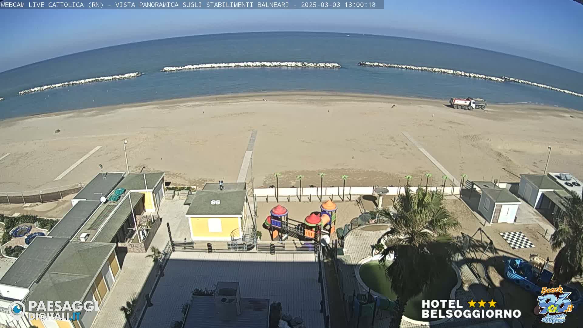 A sandy beach with calm blue water, breakwaters, beachside buildings, and a playground is seen from an elevated viewpoint on a sunny day.