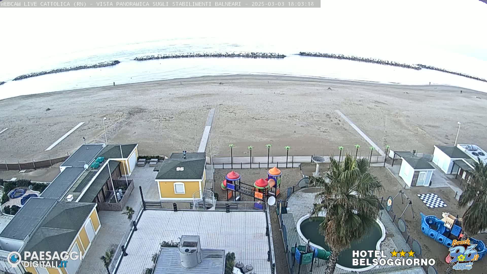 An overcast day reveals a sandy beach with calm ocean waves, breakwaters, beachside buildings, a playground, and a swimming pool.