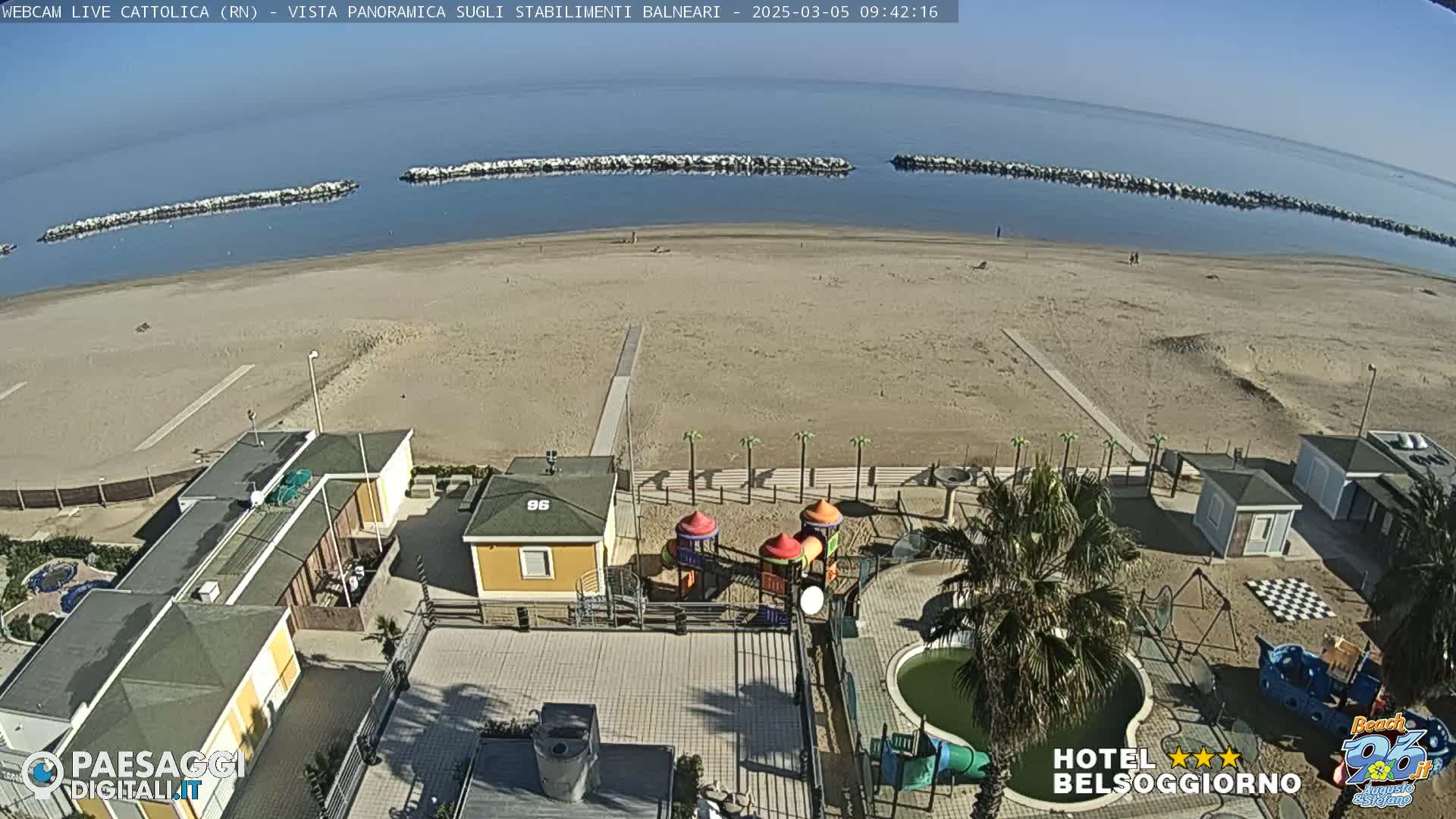 An aerial view of a sandy beach on a clear day shows beachside buildings, a playground, and calm ocean water with breakwaters.
