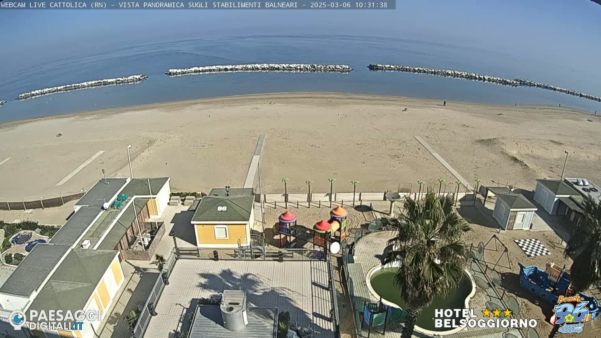 A sandy beach with calm ocean water and breakwaters, viewed from above, showing beach buildings, a playground, and palm trees on a sunny day.