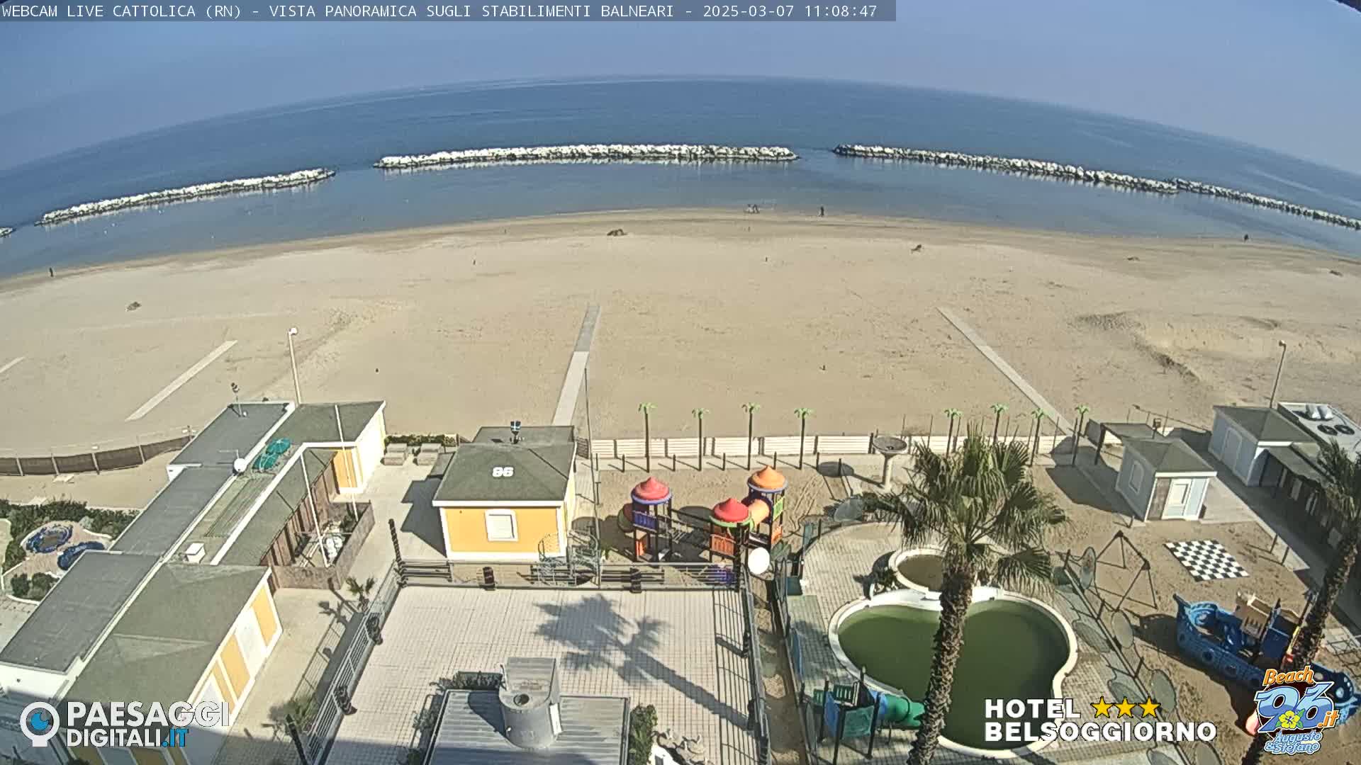 An aerial view of a sandy beach on a sunny day, showing beach structures, a playground, and a small pool.