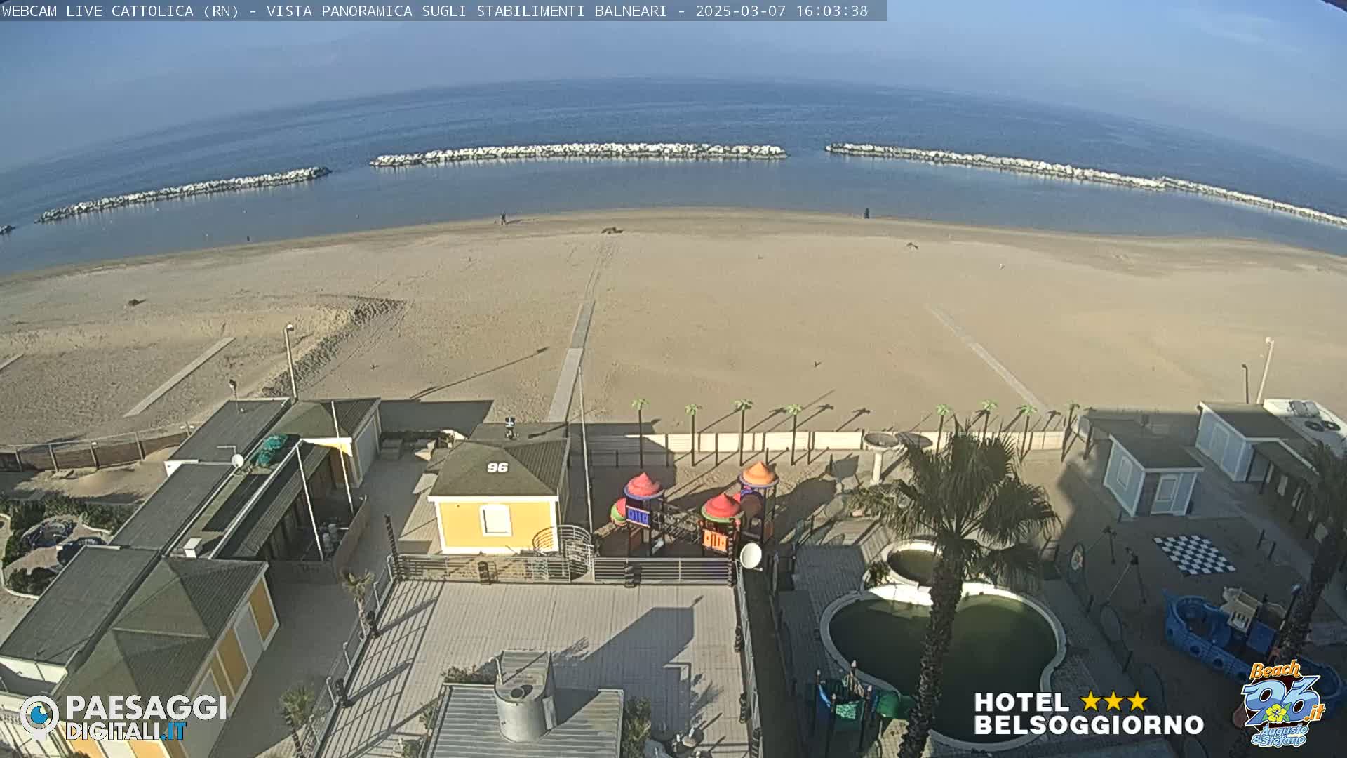 A sandy beach with calm ocean water and breakwaters, viewed from above, showing beachside buildings, a playground, and a pool on a sunny day.