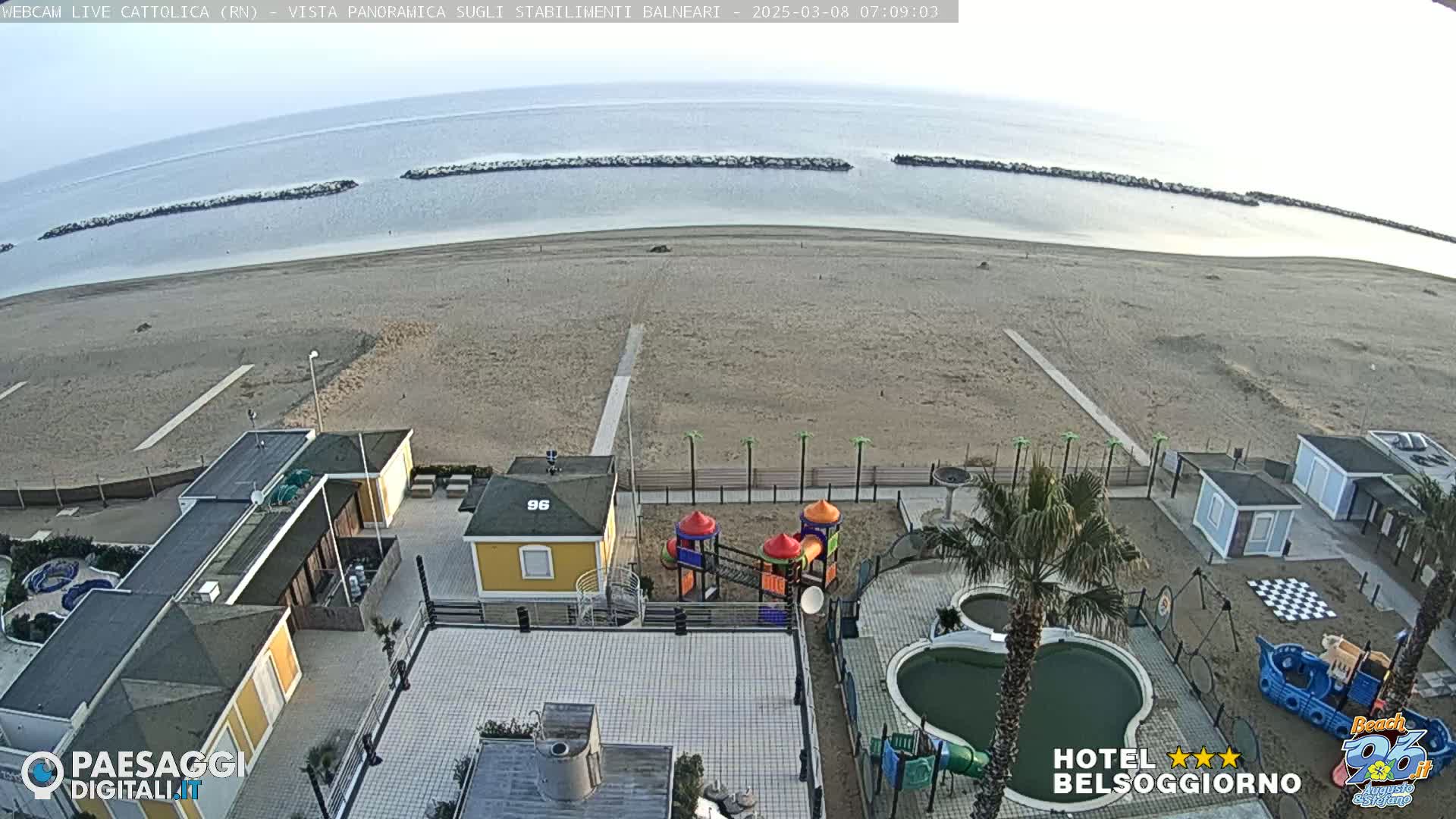 An aerial view shows a mostly empty sandy beach bordered by buildings, a playground, and a small pool, under a clear, bright sky.