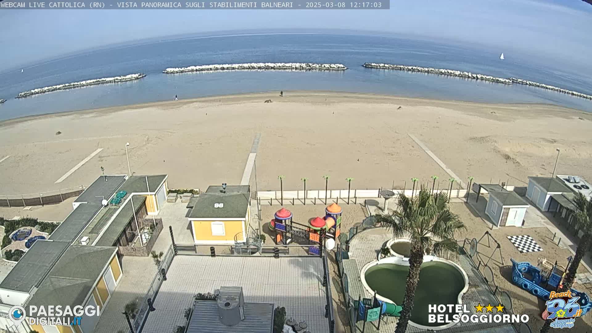 A sandy beach with calm ocean water, breakwaters, beachside buildings, a playground, and a pool are visible on a sunny day.