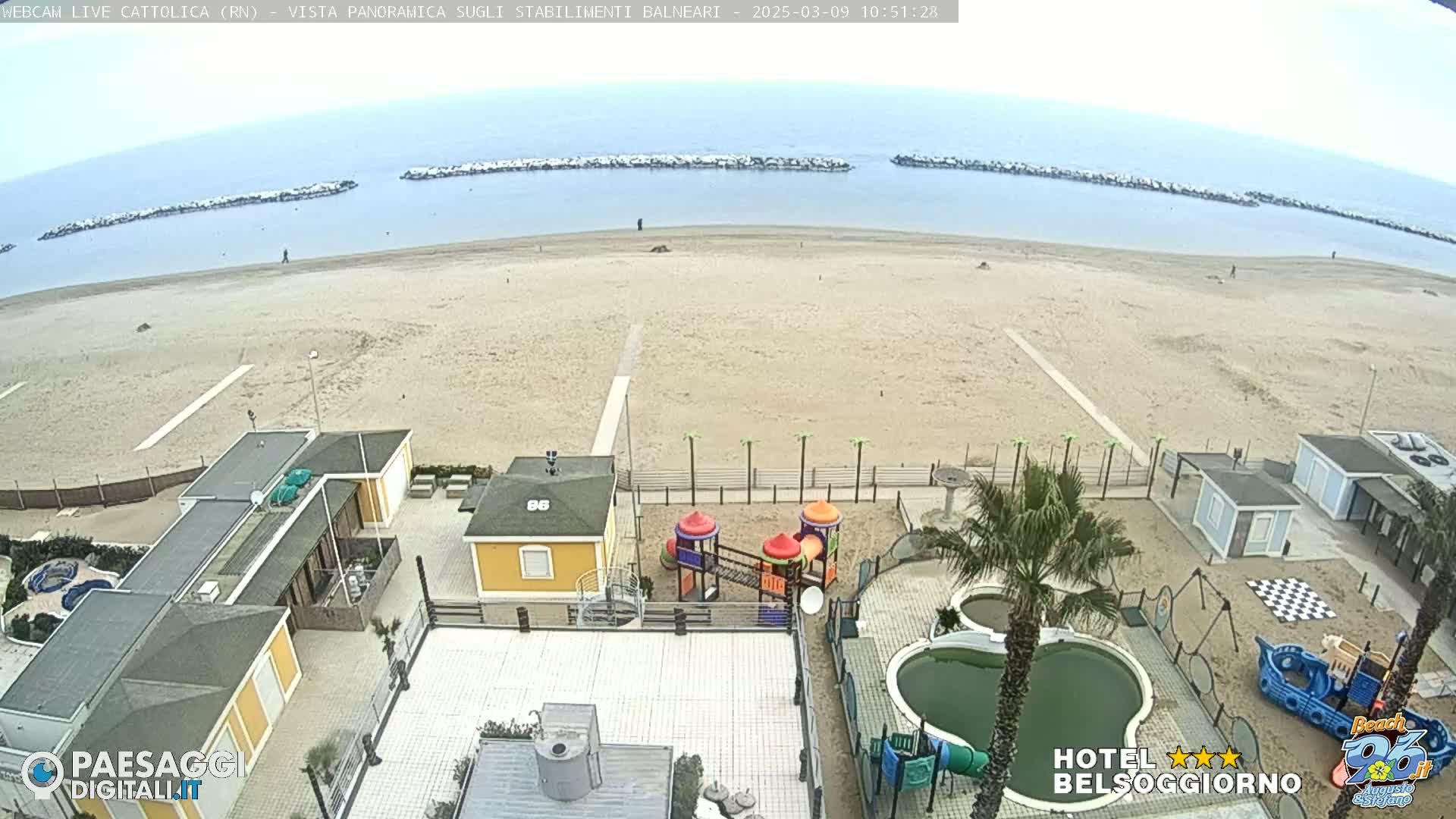 An overhead view of a sandy beach with calm, light-blue water, breakwaters, beachside buildings, a playground, and swimming pools on a mostly cloudy day.
