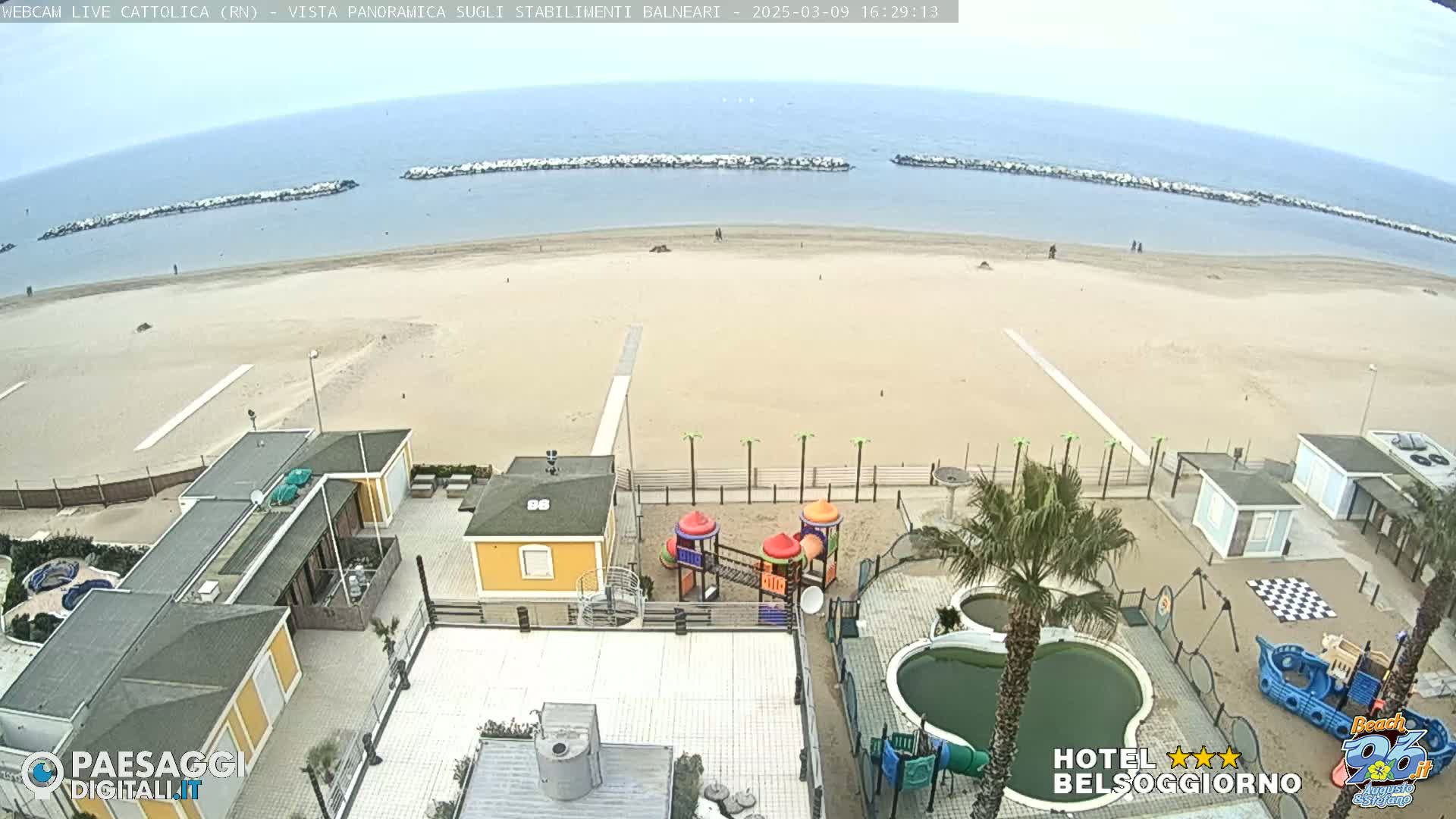 An aerial view of a sandy beach with calm ocean water, beachside buildings, a playground, and a swimming pool, under a mostly clear sky.