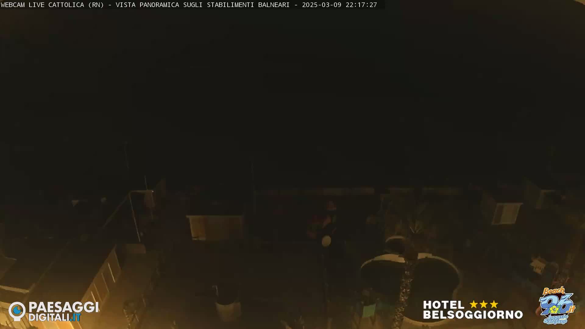 A nighttime, low-light, overhead view shows a dark area with dimly lit structures and a few indistinct figures.