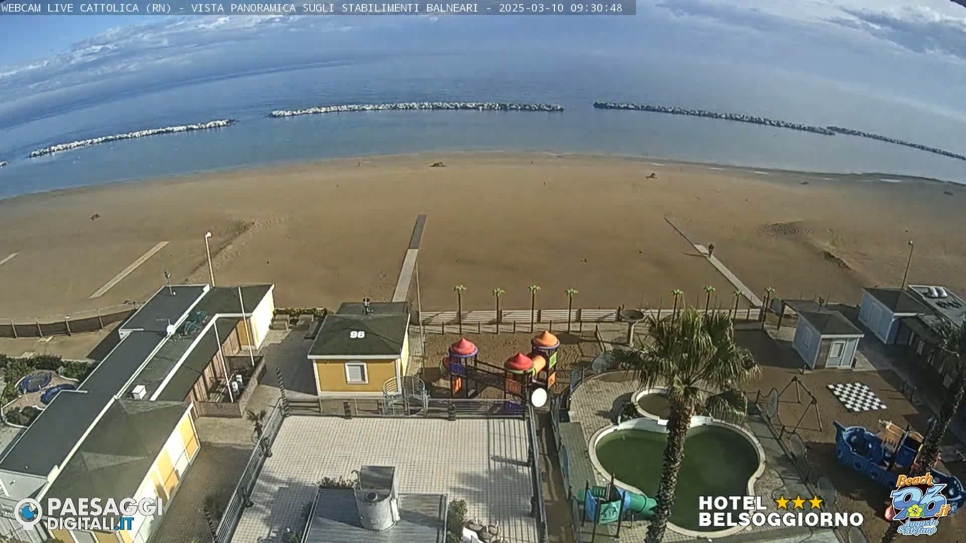 An aerial view shows a mostly empty sandy beach, calm ocean water with breakwaters, beachside buildings, a playground, and a swimming pool under a partly cloudy sky.