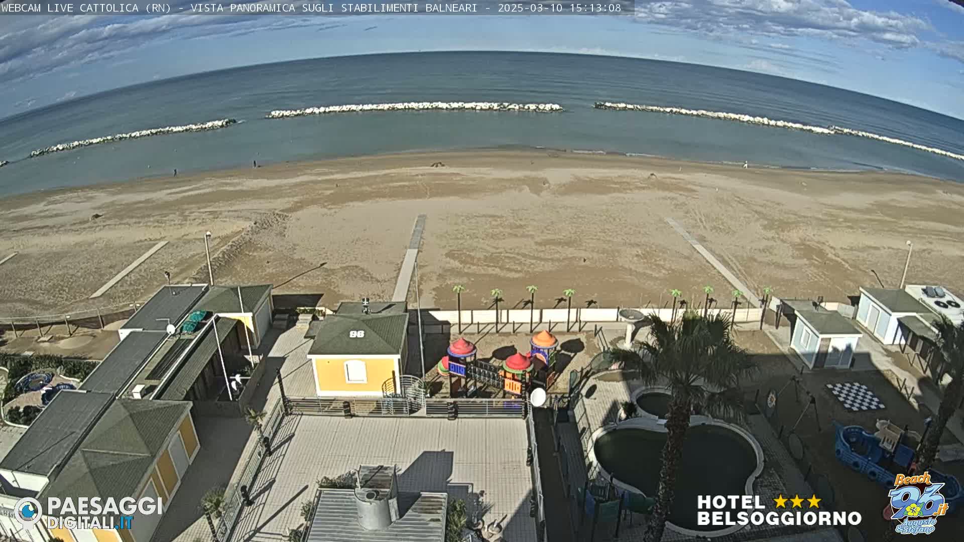 An aerial view shows a sandy beach with calm ocean waters under a partly cloudy sky, alongside beachside buildings, a playground, and swimming pools.