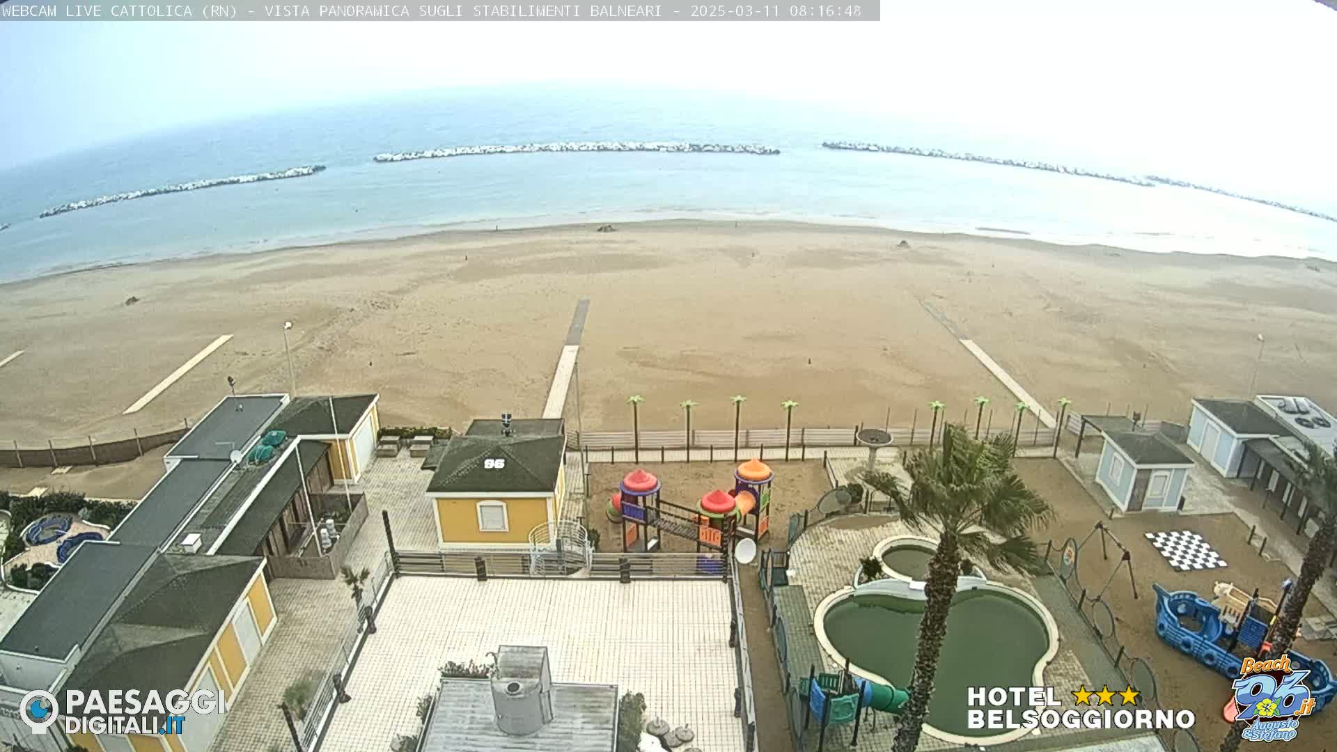 An overcast day at a sandy beach with calm water, beachside buildings, a playground, and pools.