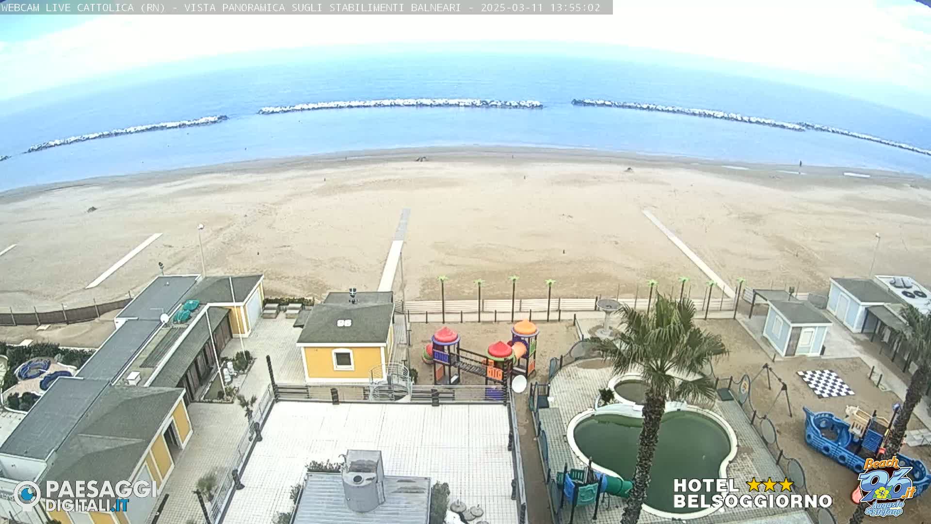 An aerial view shows a mostly empty sandy beach with calm ocean water, beachside buildings, a playground, and swimming pools, under a partly cloudy sky.