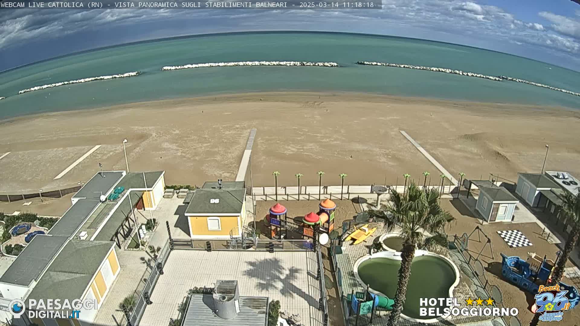 A sandy beach stretches to a calm, teal ocean under a partly cloudy sky, overlooking a beachfront area with buildings, a playground, and small pools.