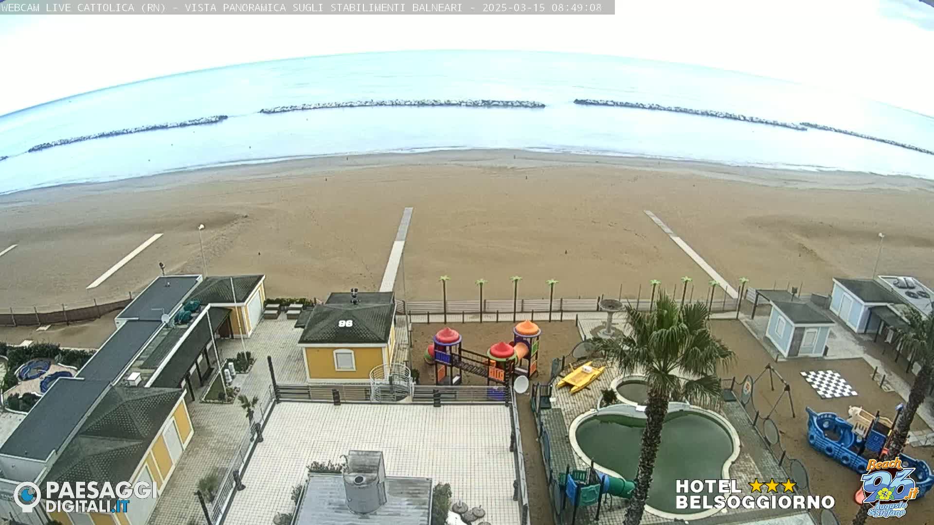 A sandy beach with calm, light-blue water under an overcast sky is viewed from above, showing beachside buildings, a playground, and swimming pools.
