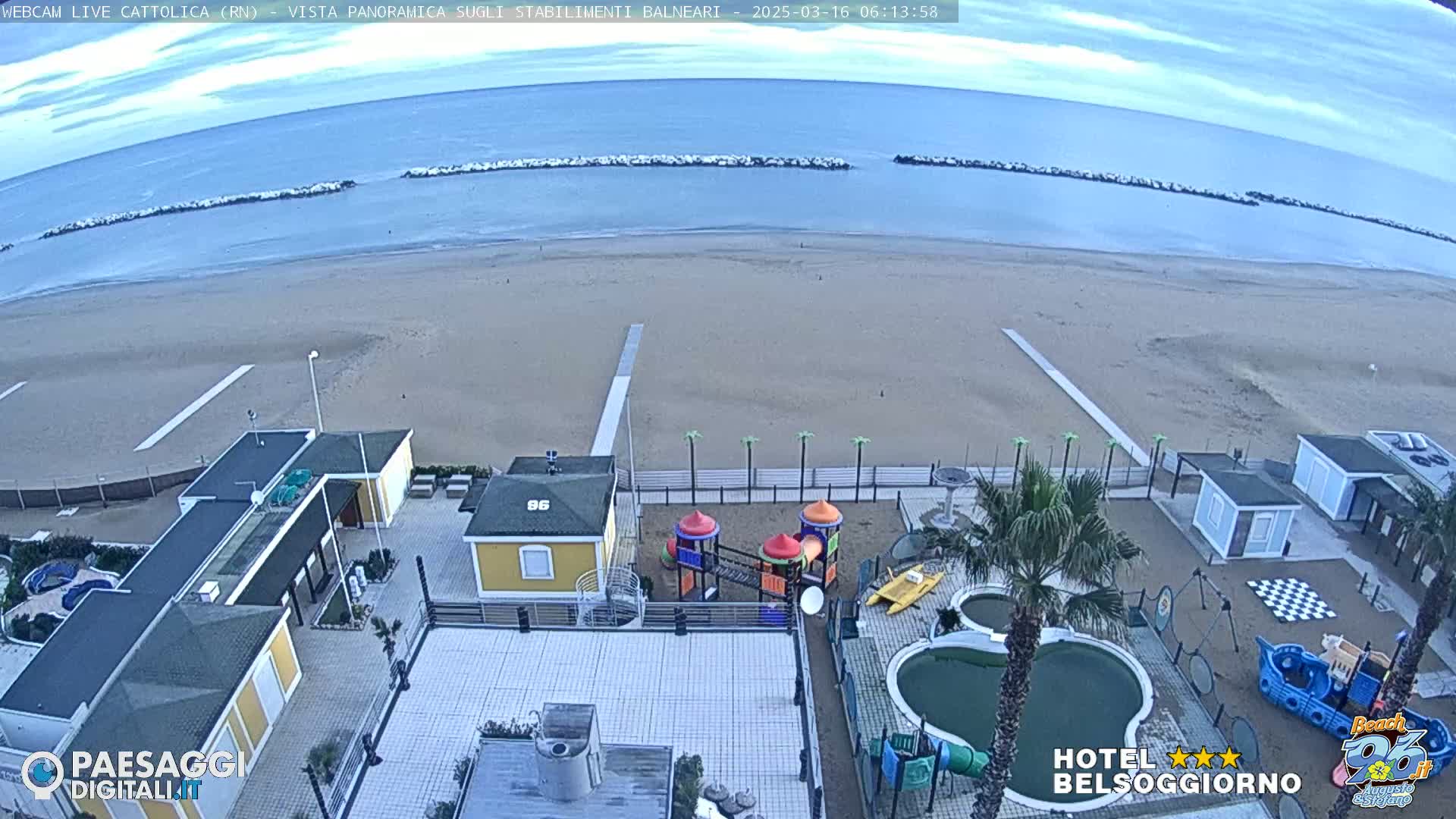 An aerial view of a sandy beach with calm ocean water and a resort area including a playground, swimming pool, and small buildings, under an overcast sky.