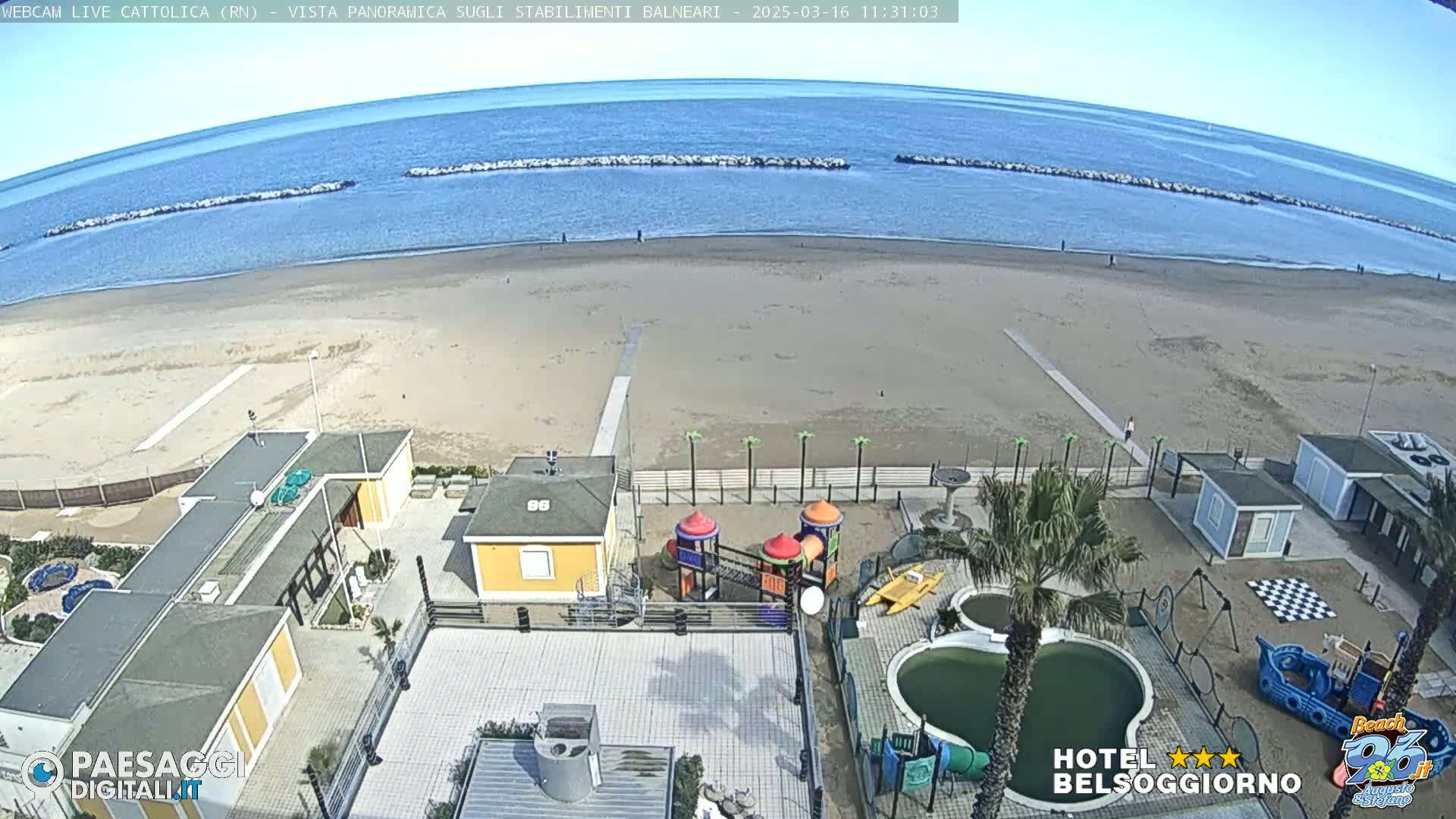 A sandy beach with calm blue water and a breakwater is visible from an elevated viewpoint overlooking beachside buildings, a playground, and a swimming pool.