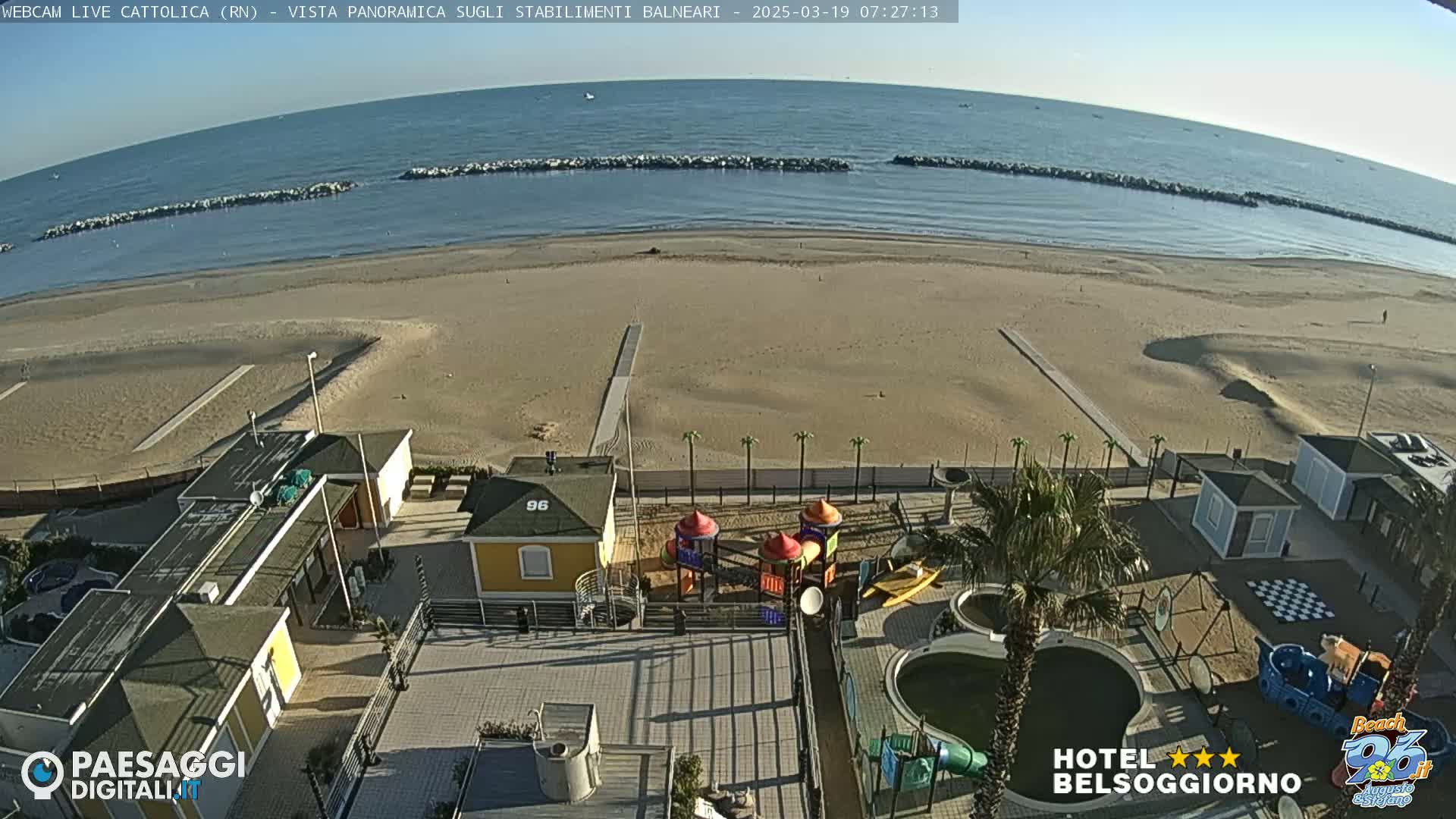 A beach with calm ocean water under a clear sky is viewed from above, showing beachside buildings, a playground, and a pool area.