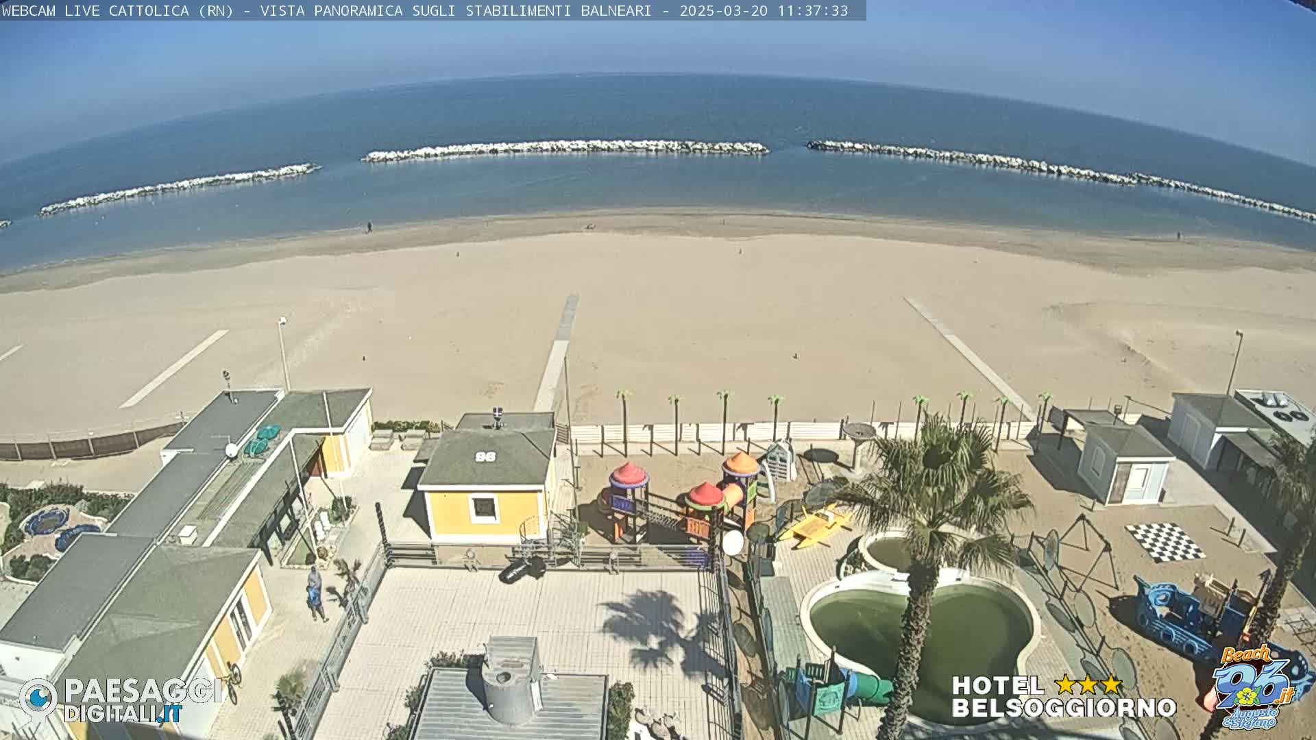 A beach with a playground and buildings is shown under a clear, sunny sky; the ocean is calm.