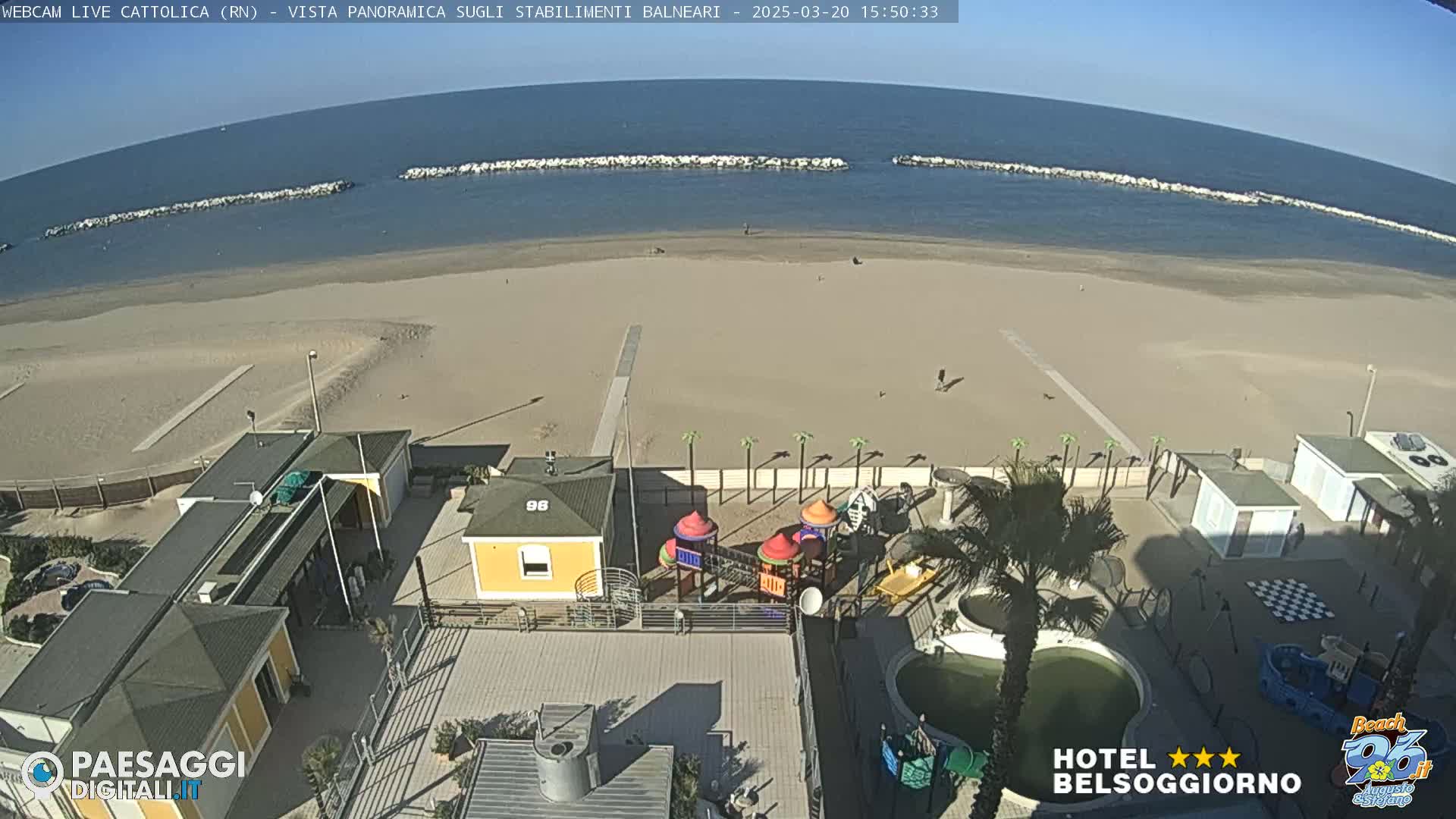 A beach scene on a sunny day shows a sandy beach with calm water, a breakwater, beach buildings, a playground, and palm trees.