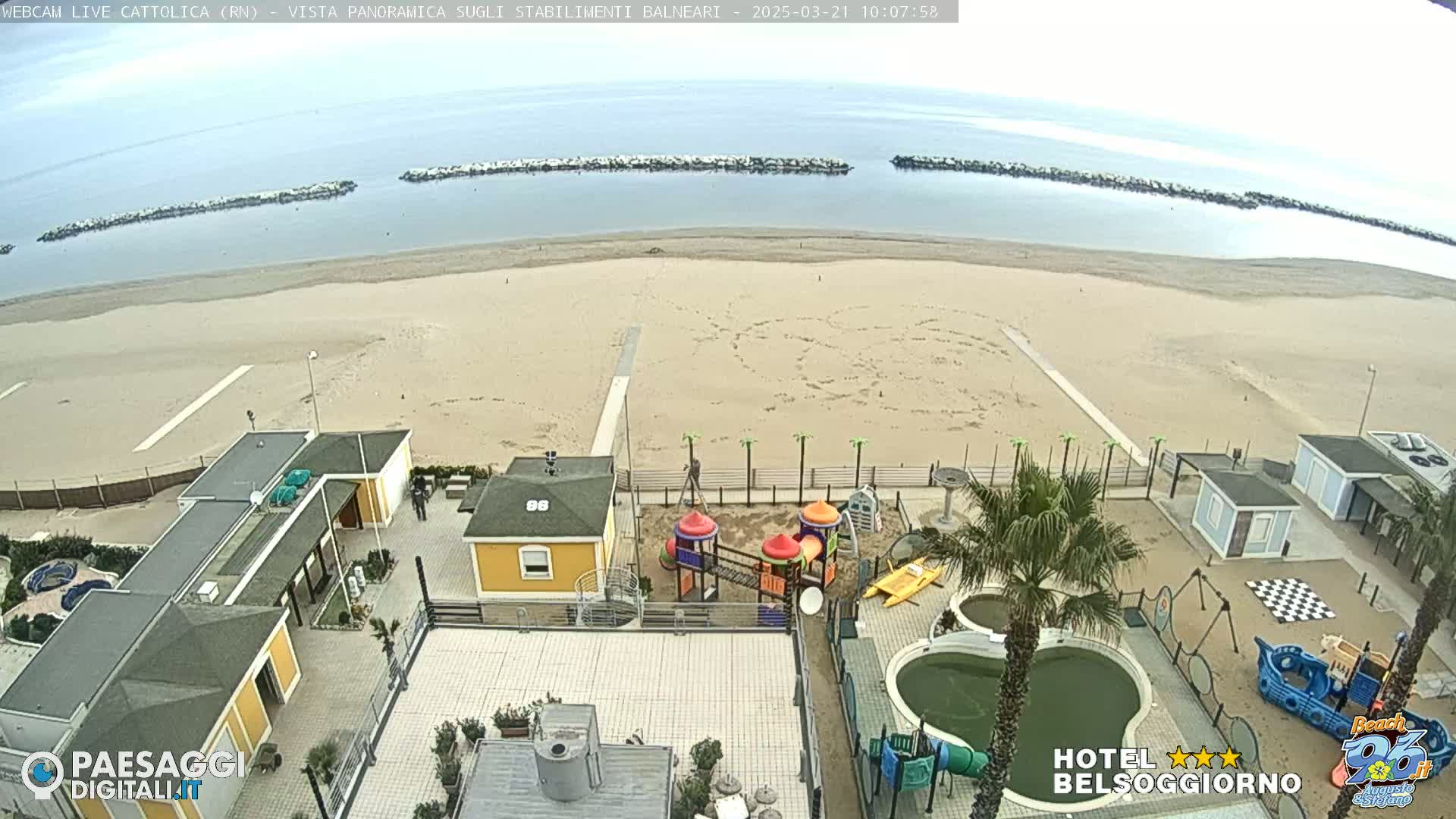 An aerial view shows a sandy beach with calm, light-blue water, breakwaters, and a playground and pool area at a beachfront hotel complex under an overcast sky.