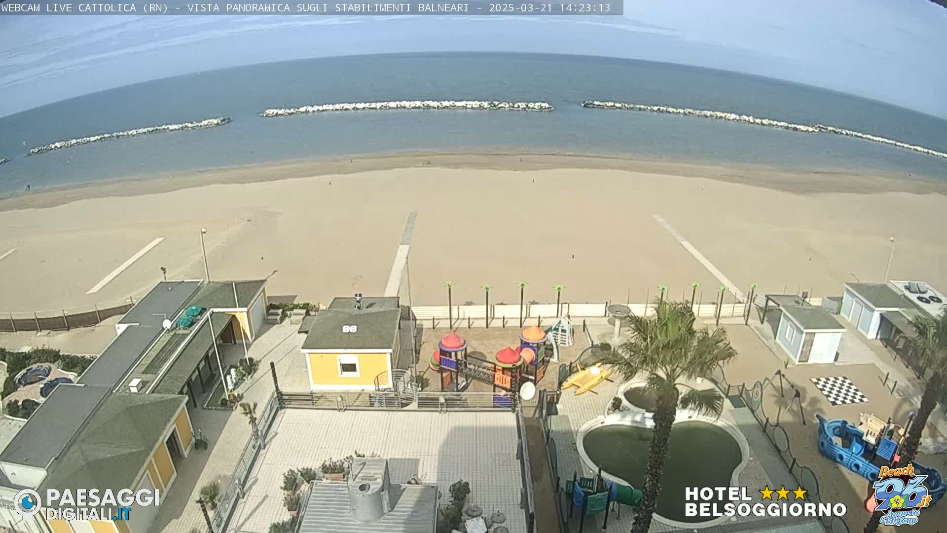 A sandy beach stretches to a calm sea, protected by a long breakwater, under a clear sky; a playground and swimming pool are visible near beachfront buildings.