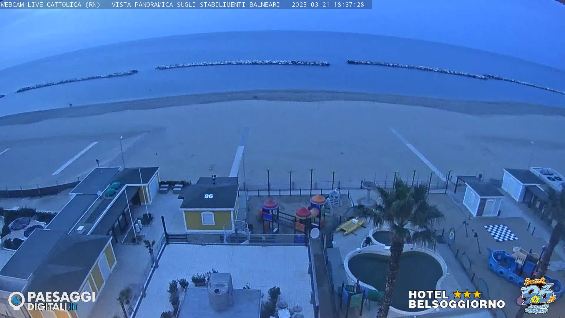 A mostly empty beach with calm water under an overcast sky is viewed from above, showing beachside buildings, a playground, and a pool.
