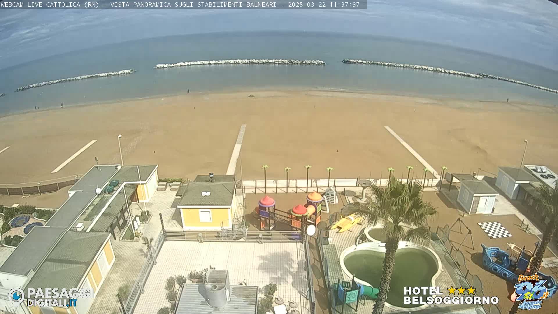 A sandy beach with calm, light blue water under a partly cloudy sky, featuring beachside buildings, a playground, and swimming pools.