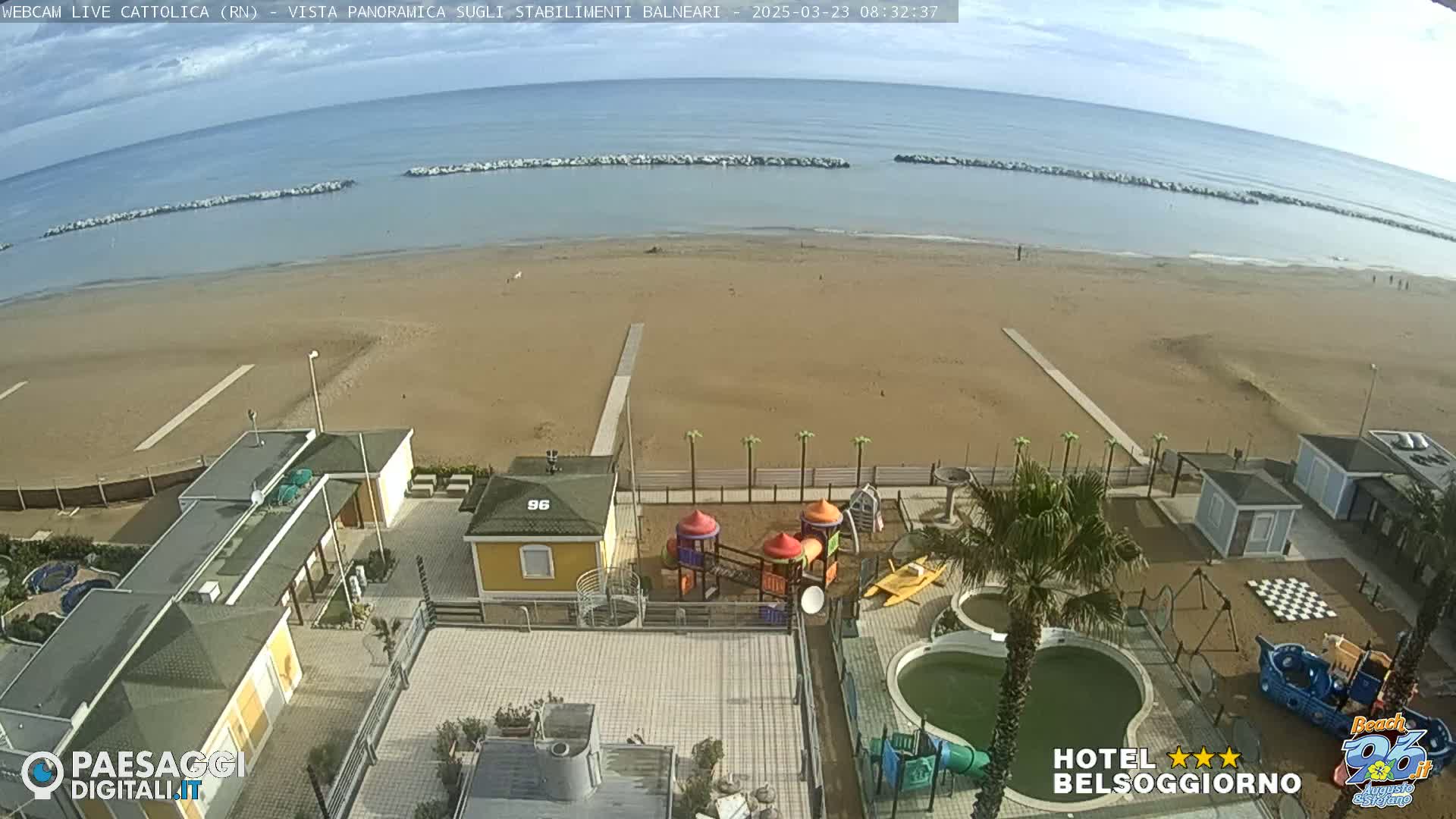 A sandy beach with calm ocean waters under a partly cloudy sky is overlooked by beachside buildings, a playground, and a small pool.
