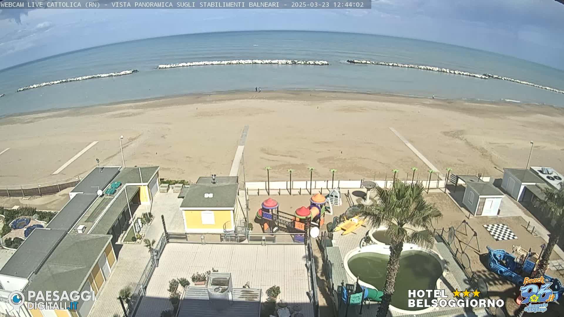 A sandy beach with calm ocean under a mostly sunny sky is viewed from above, showing beach buildings, a playground, and a small pool.