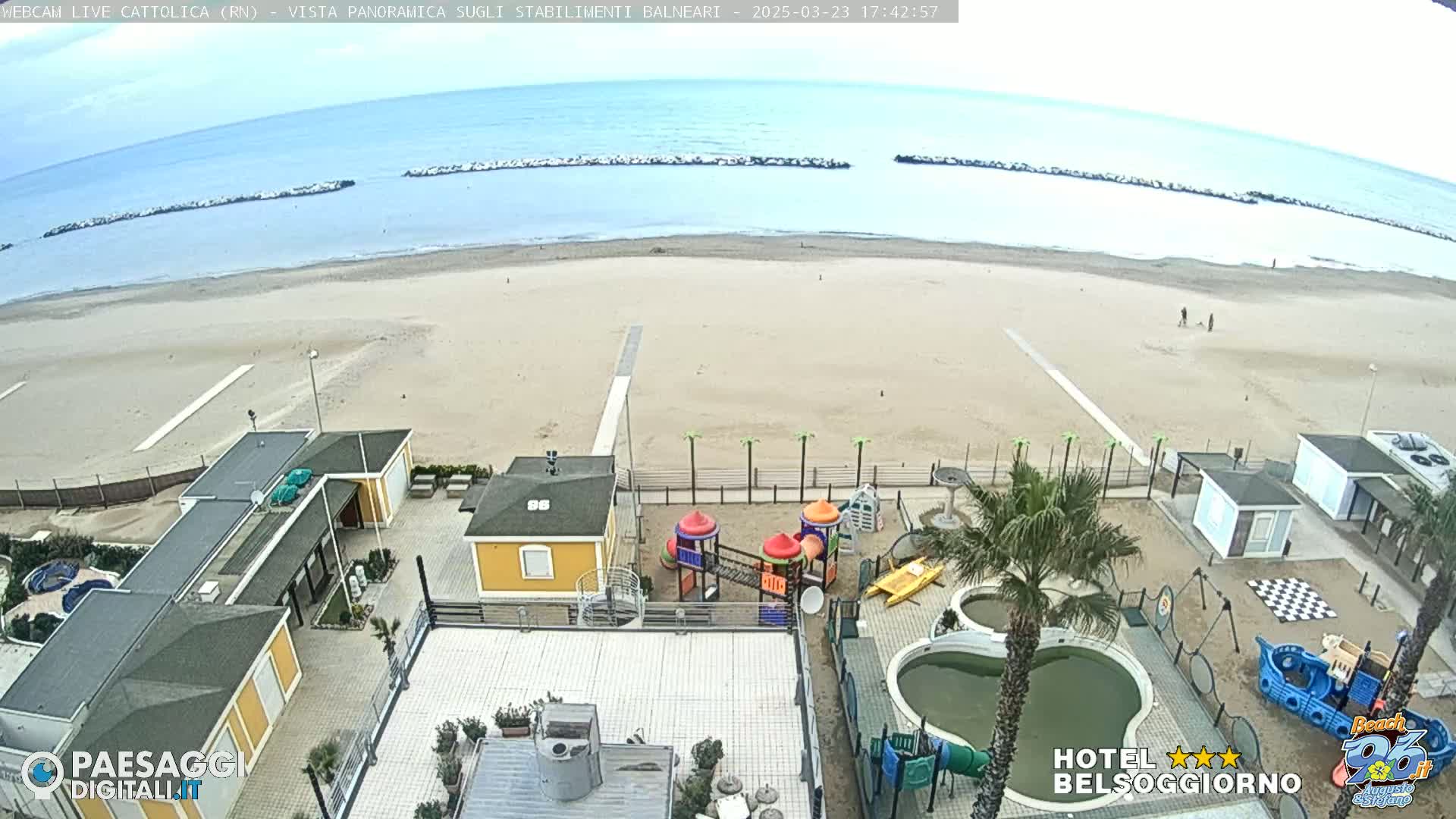 An aerial view of a beach with calm water under a mostly clear sky shows beach buildings, a playground, and several small pools.