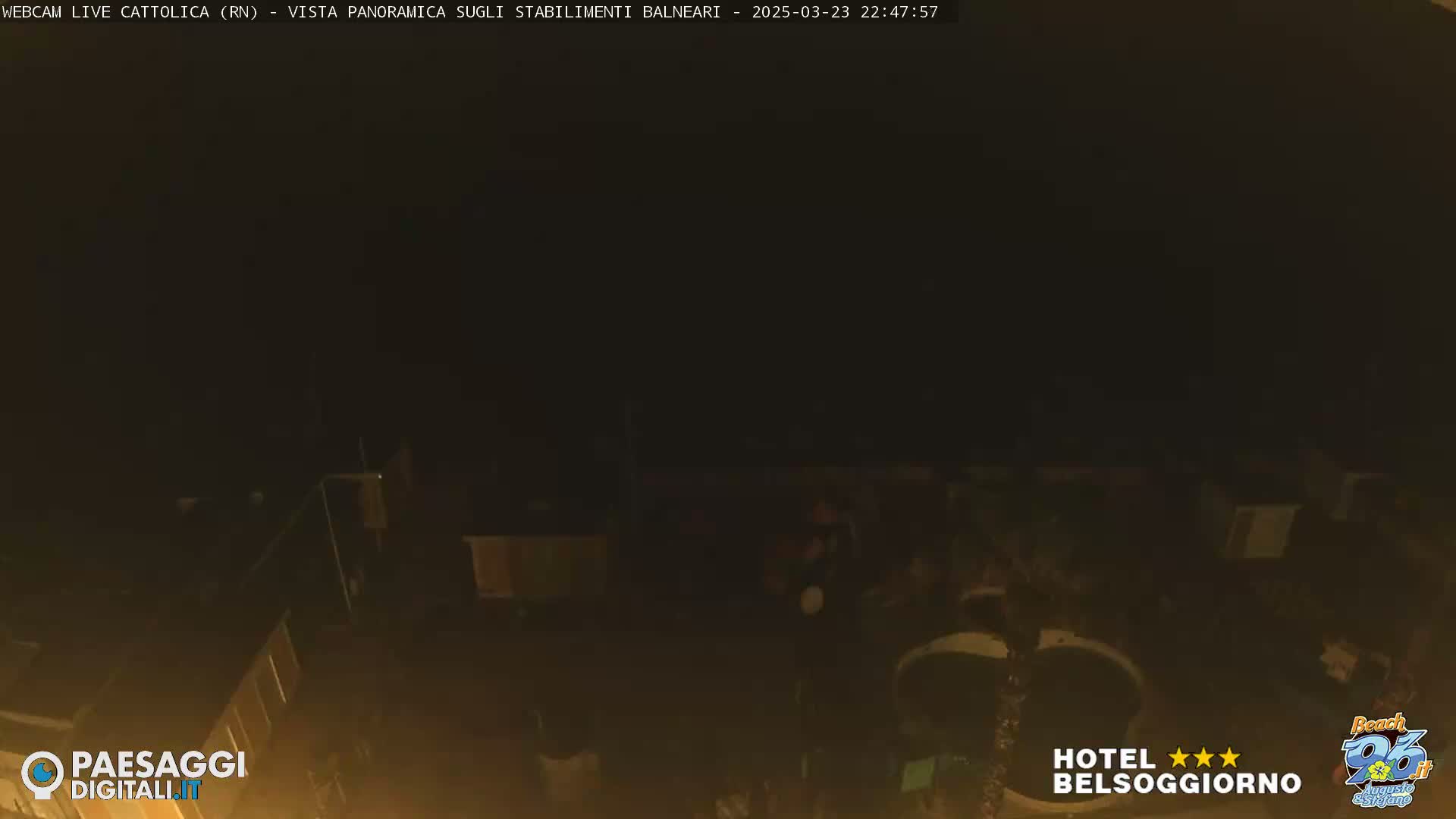 A dark, nighttime view of a beach area with dimly lit structures and a few indistinct figures.