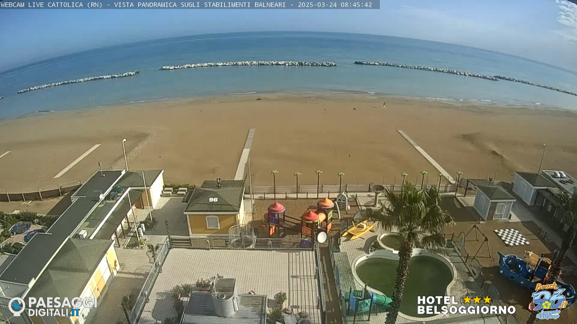 A mostly sunny beach scene shows a sandy beach with calm water, beachside buildings, a playground, and a small pool.