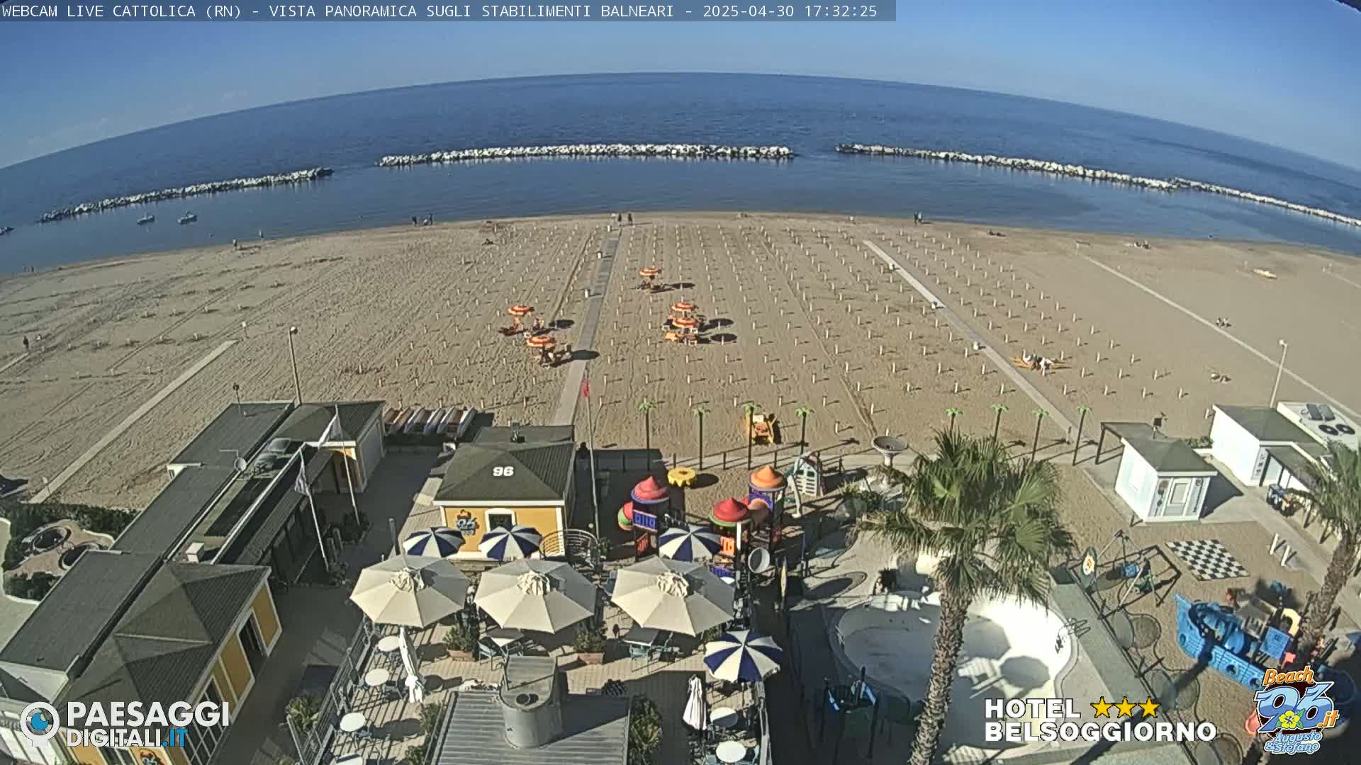 A mostly empty sandy beach with rows of umbrellas and a playground is situated next to a calm ocean under a clear sky.