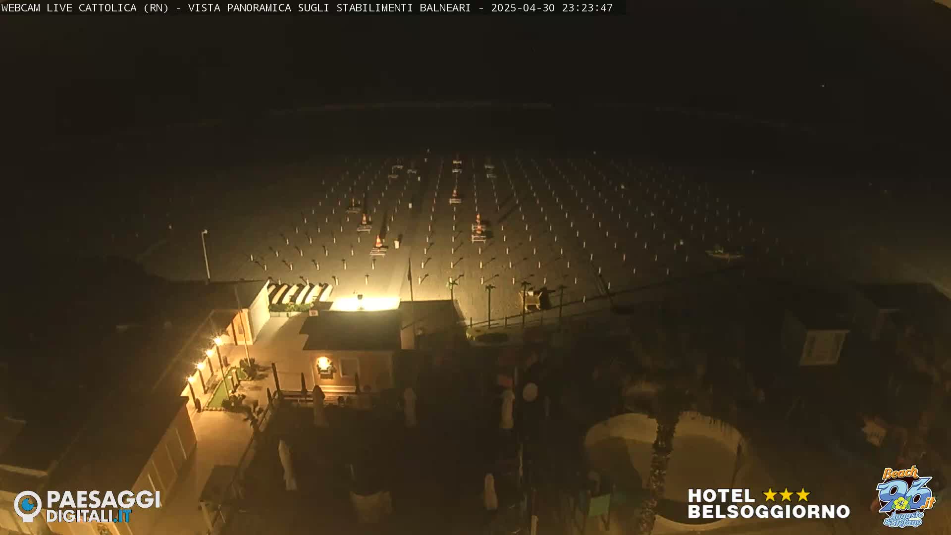 A nighttime, aerial view of a beach with rows of empty beach chairs and umbrellas, and a building illuminated with lights.