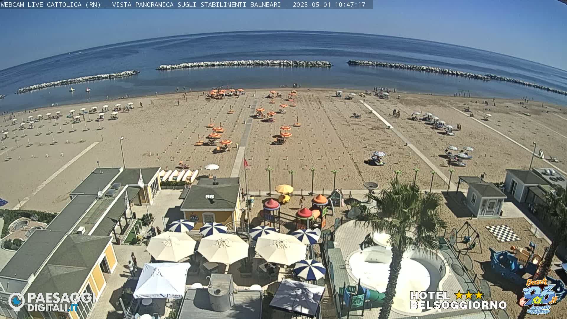 A sandy beach with numerous umbrellas and beachgoers is shown, adjacent to a beachfront area with buildings, playground equipment, and a small pool; the weather is sunny and clear.