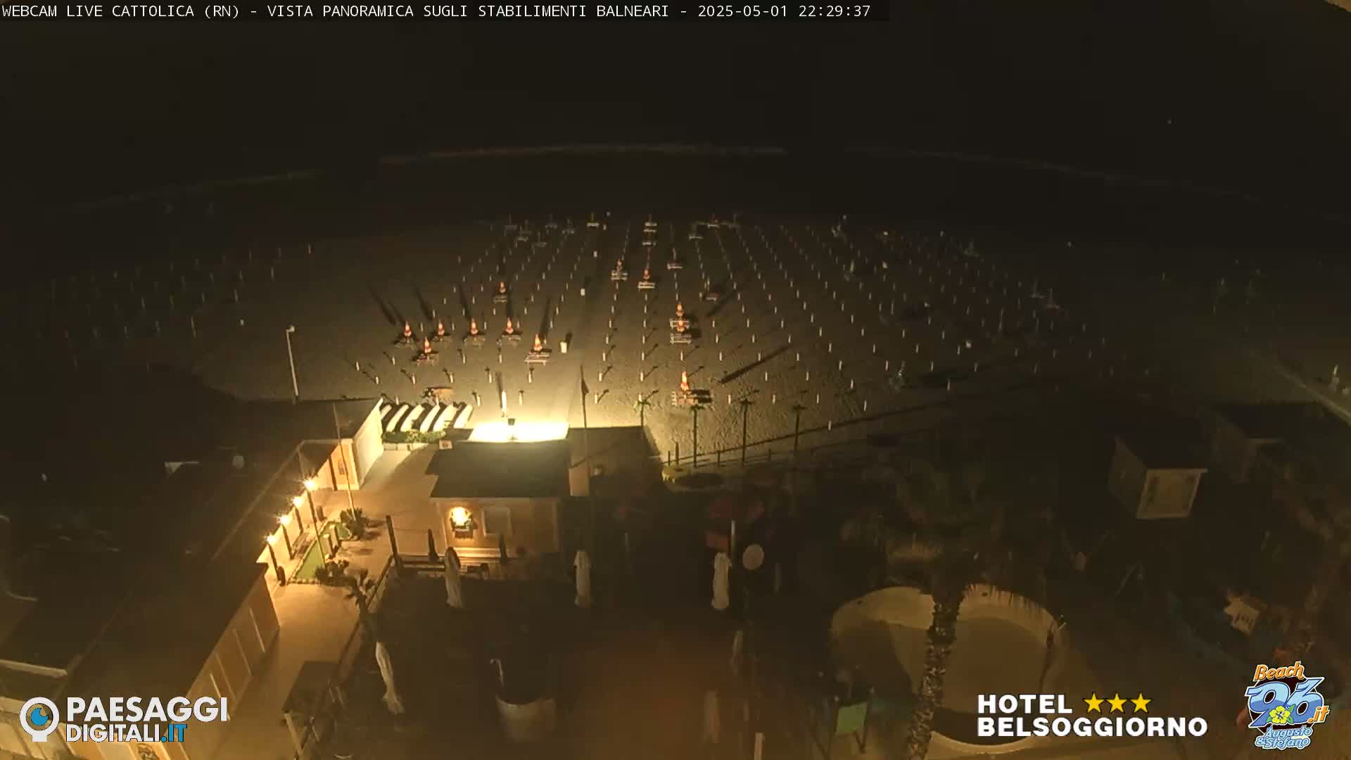 A nighttime, aerial view of a beach at night shows numerous empty beach umbrellas and chairs arranged in rows, along with a building with lights on near the shore.