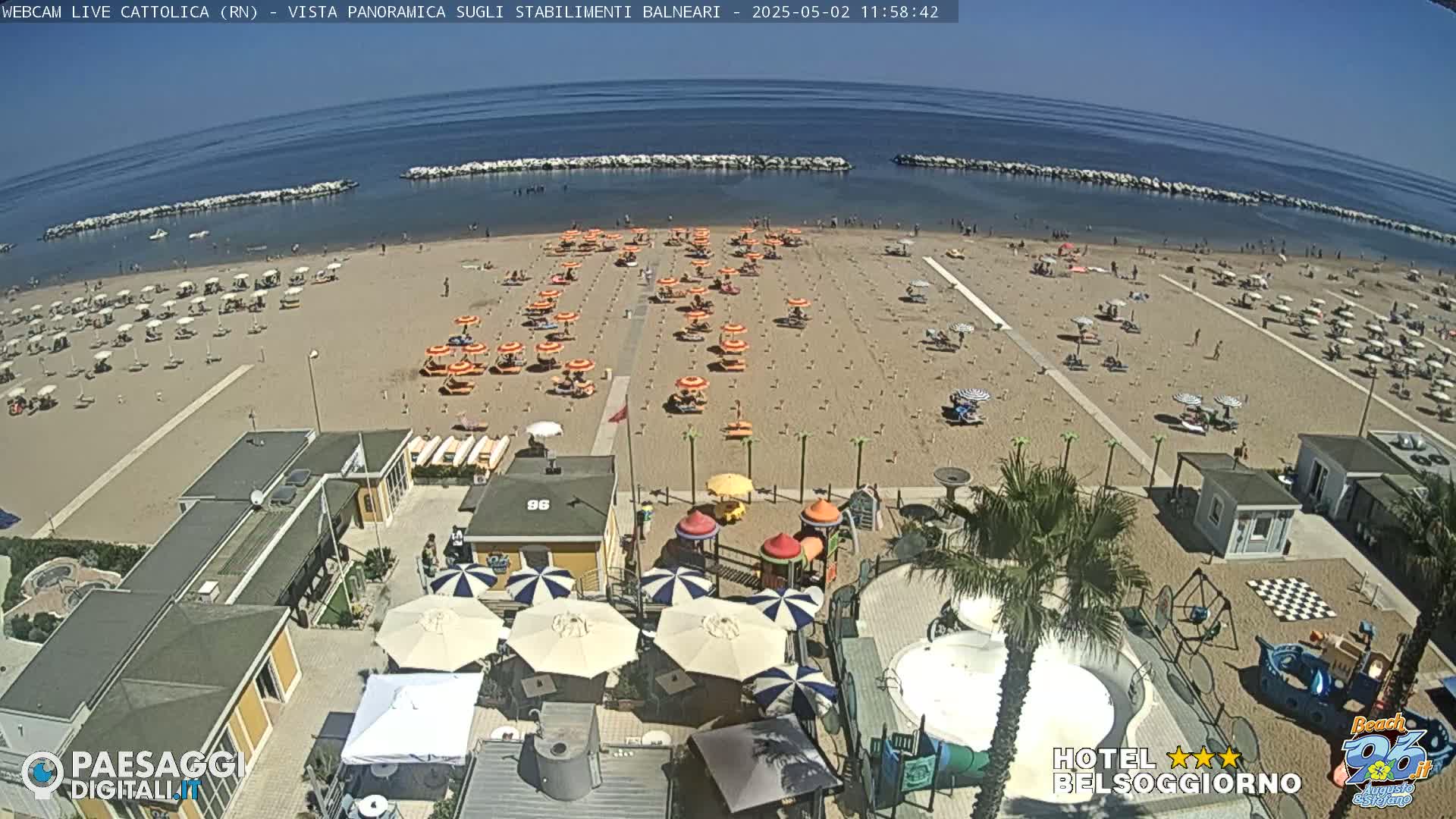 A sandy beach with numerous umbrellas and beachgoers is seen from an elevated viewpoint on a sunny day, alongside beachside buildings and a children's play area.
