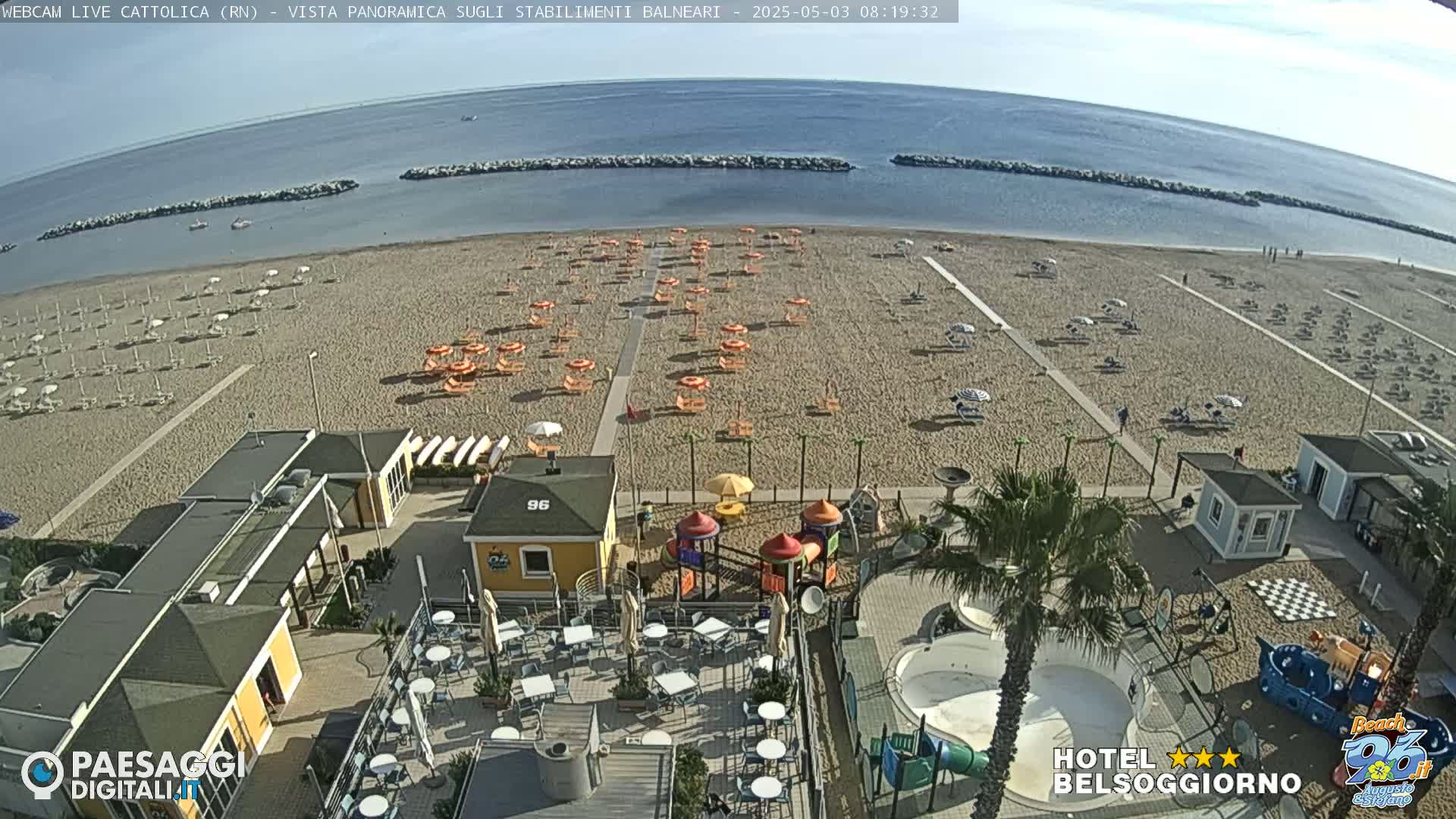 A sandy beach with numerous orange and white umbrellas, a playground, and beachside buildings is visible under a clear sky.