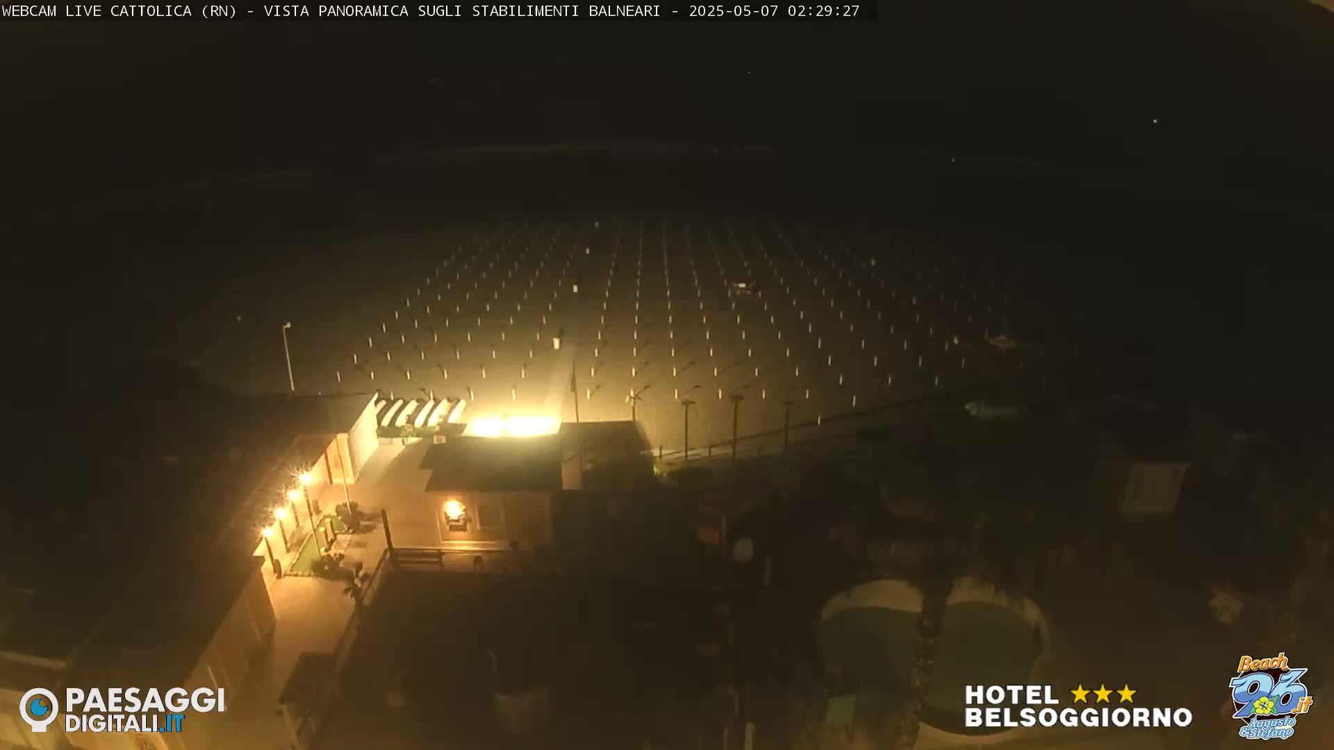 A dark, nighttime view of an empty beach with rows of beach umbrellas and a building illuminated by lights.