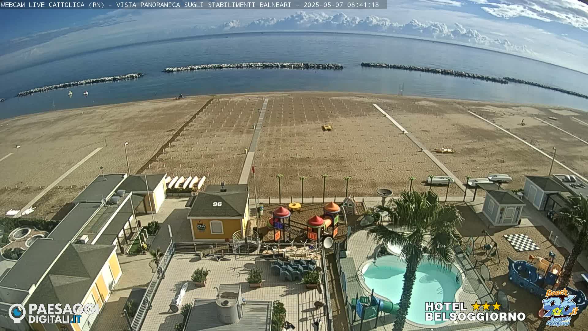 A beach with calm ocean, beach umbrellas, and a playground, viewed from an elevated position overlooking buildings with a swimming pool.