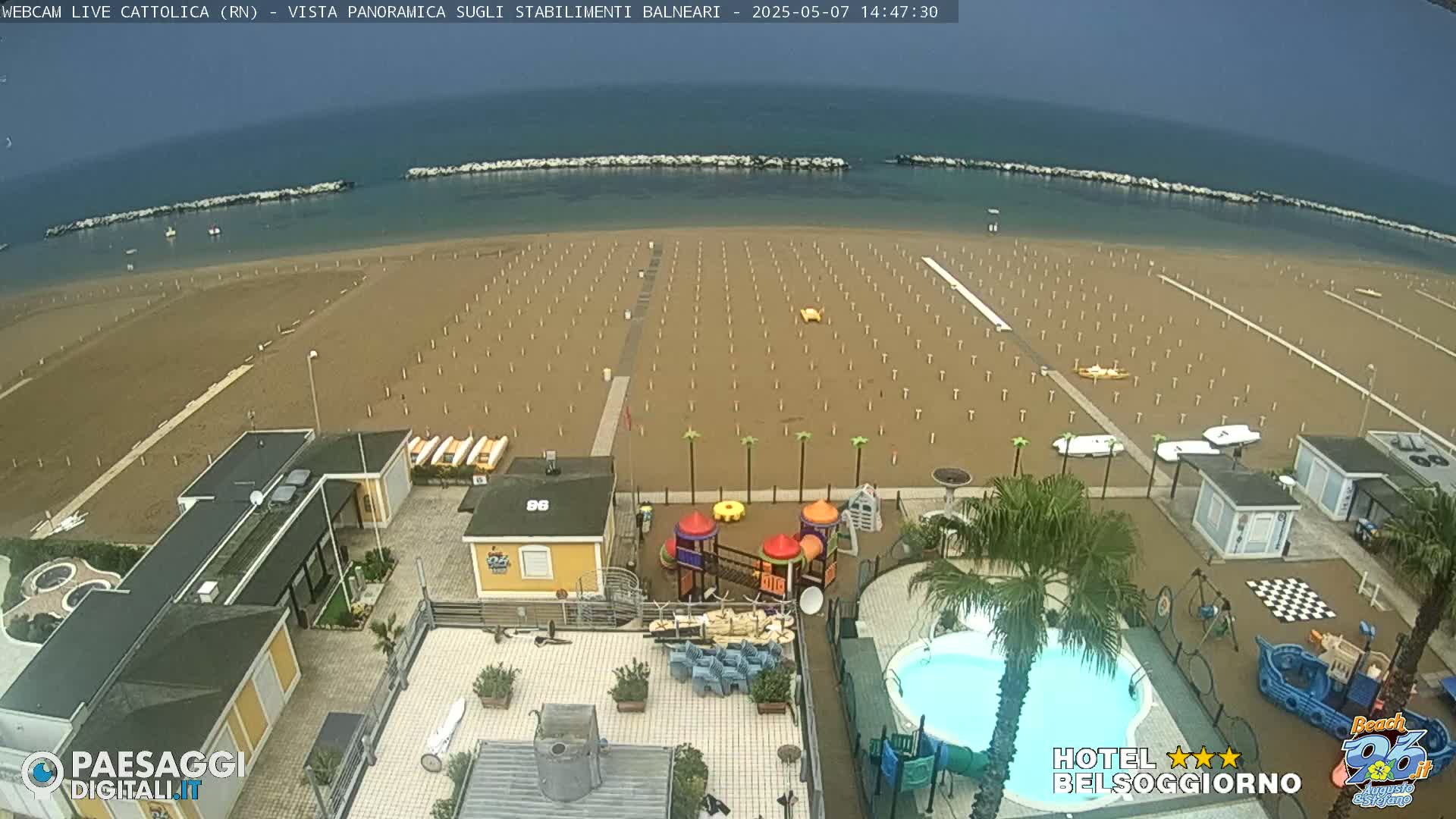 An overcast day reveals a beach with rows of empty beach umbrellas, a swimming pool, and a playground at a beachfront hotel.