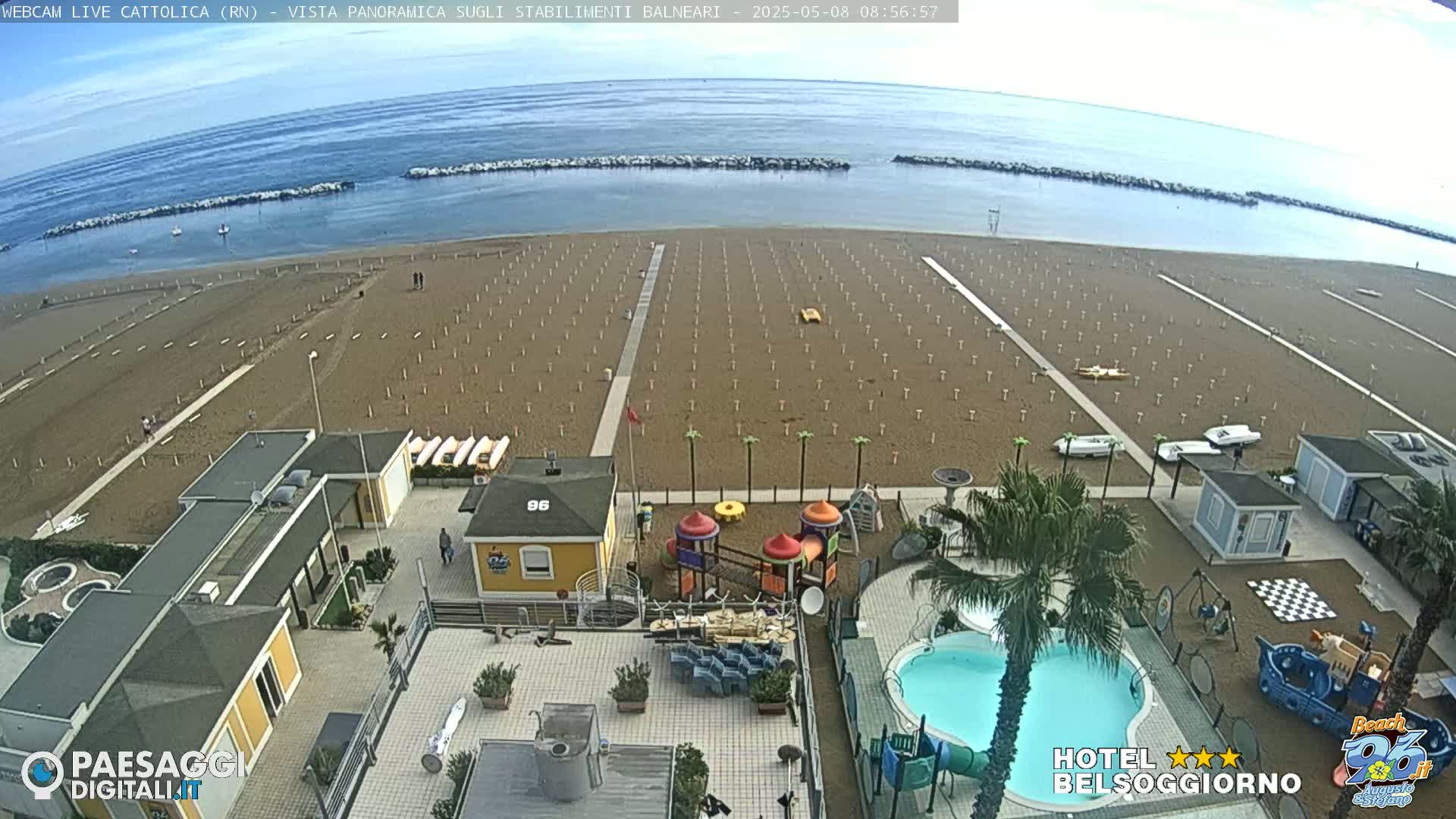 An empty sandy beach with rows of unoccupied beach umbrellas and lounge chairs, a small playground, a swimming pool, and beachside buildings is visible on a partly cloudy day.