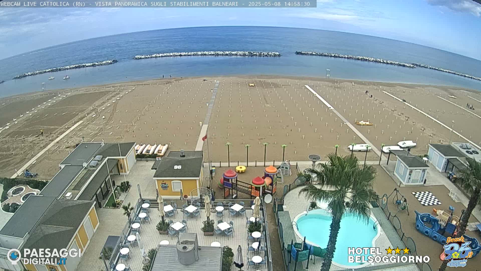 An aerial view shows a sandy beach with rows of empty umbrellas and beach chairs, a calm blue ocean, and a beachfront hotel with a pool and playground.
