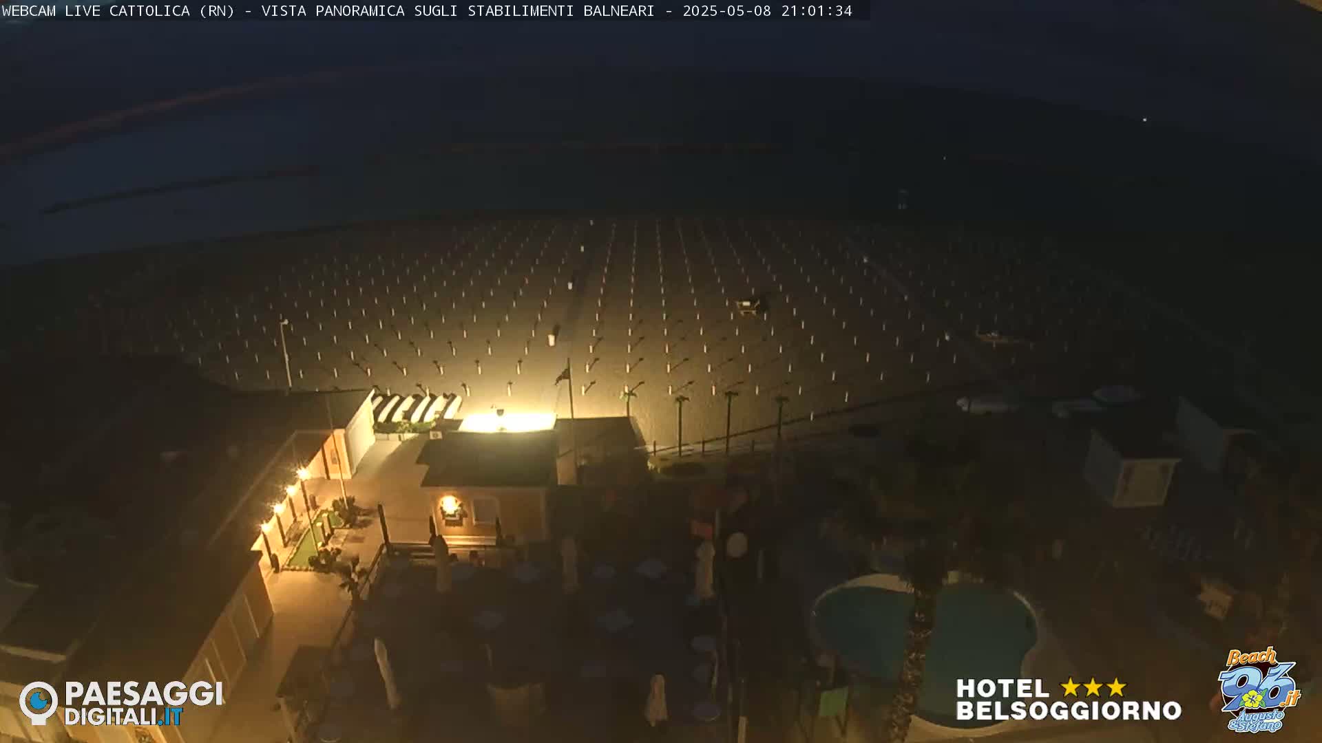 An empty beach at night with rows of beach umbrellas and a building with lights on, near a pool.