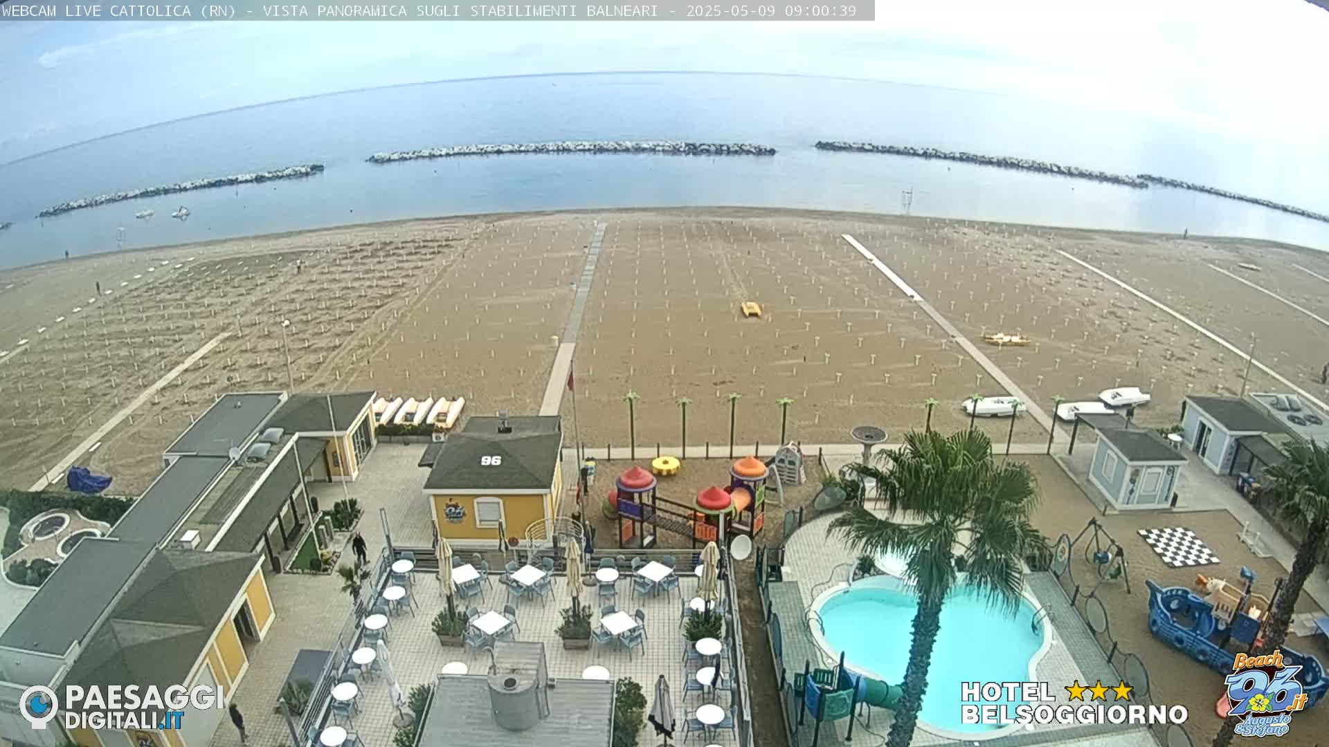 An overcast day at a beachside resort shows a mostly empty sandy beach with rows of empty beach umbrellas, a swimming pool, and playground equipment near several buildings.