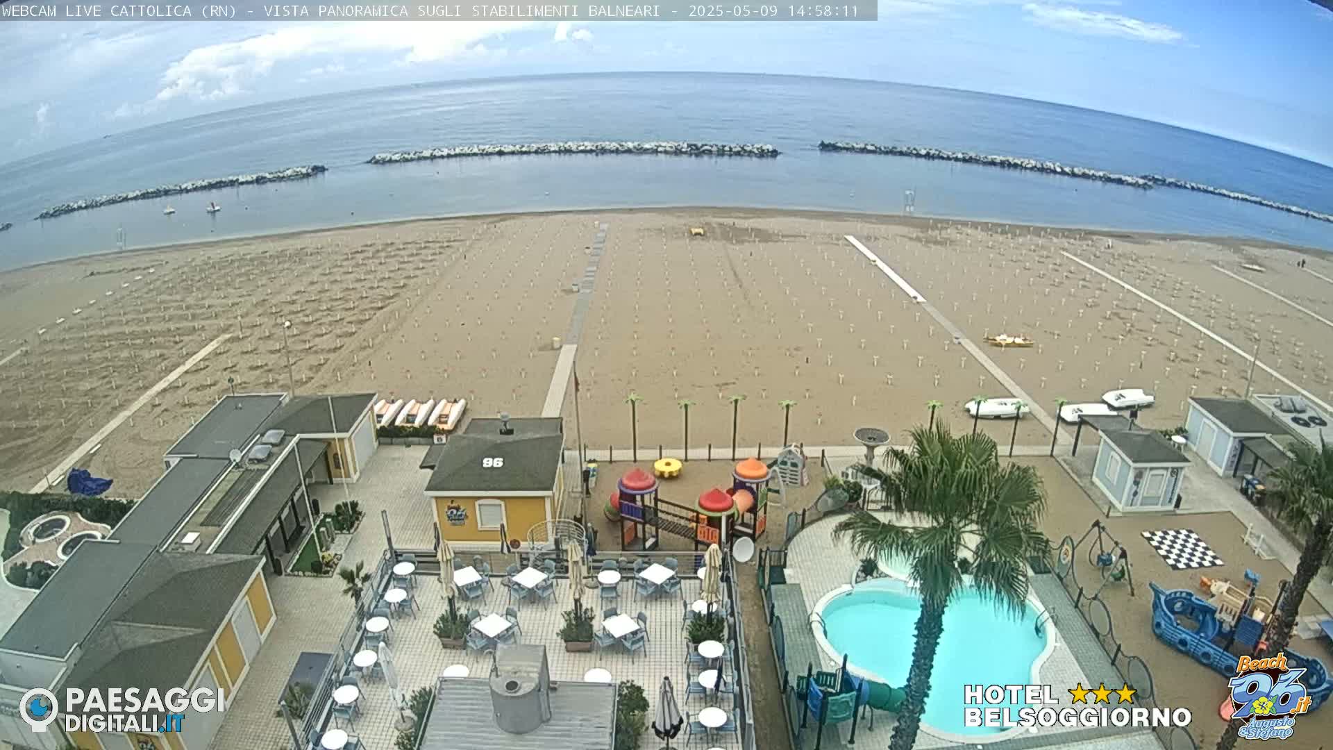 An aerial view shows a mostly empty sandy beach with rows of umbrellas, a calm sea, and a beachfront hotel with a pool and playground.
