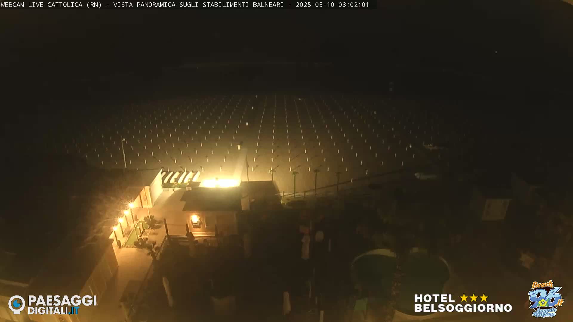 An aerial nighttime view of a beach with rows of beach umbrellas illuminated, a small building with lights on, and a few people near it.