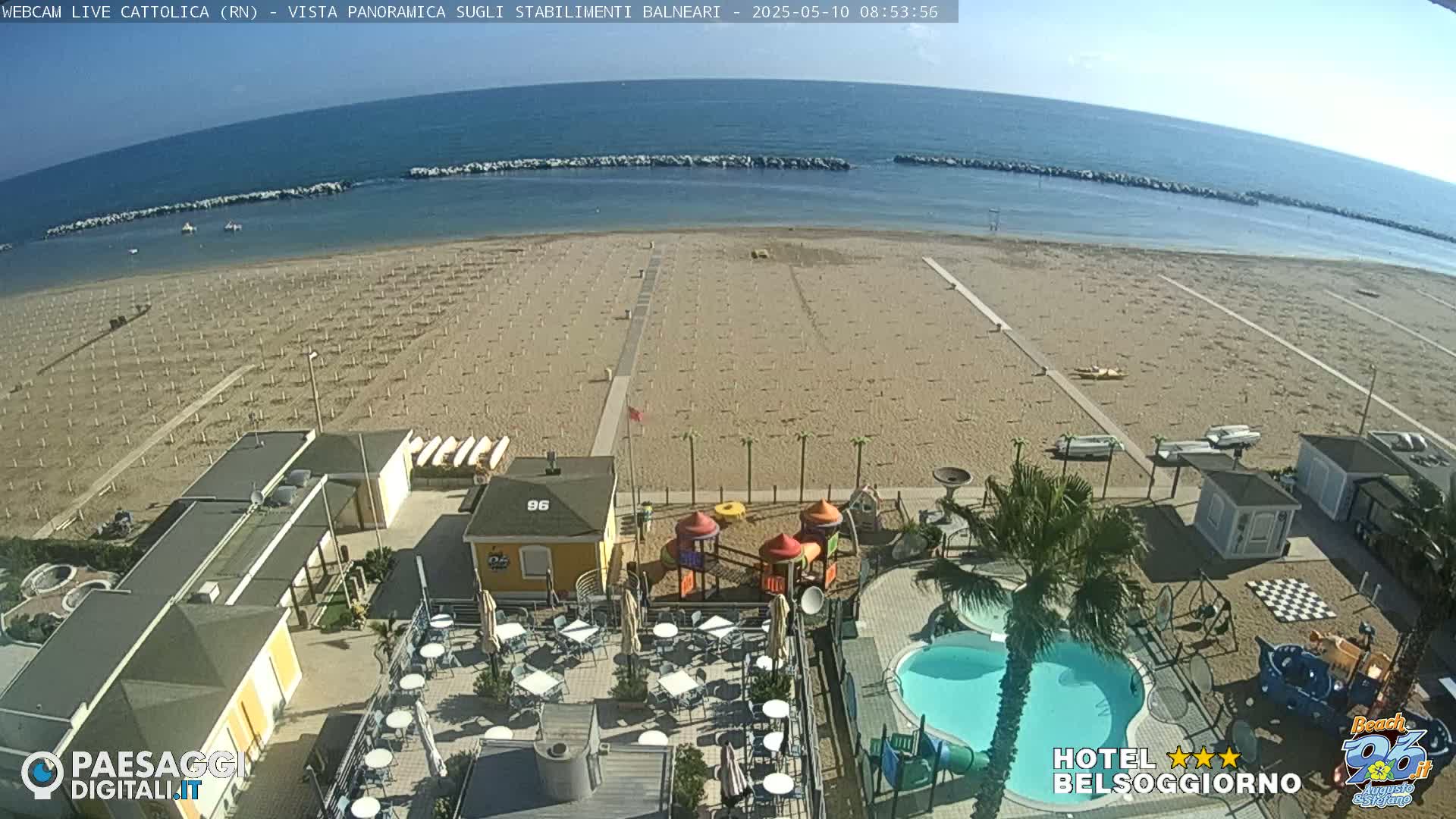 An aerial view of a beach with numerous beach umbrellas, a playground, a swimming pool, and beachside buildings on a sunny day.