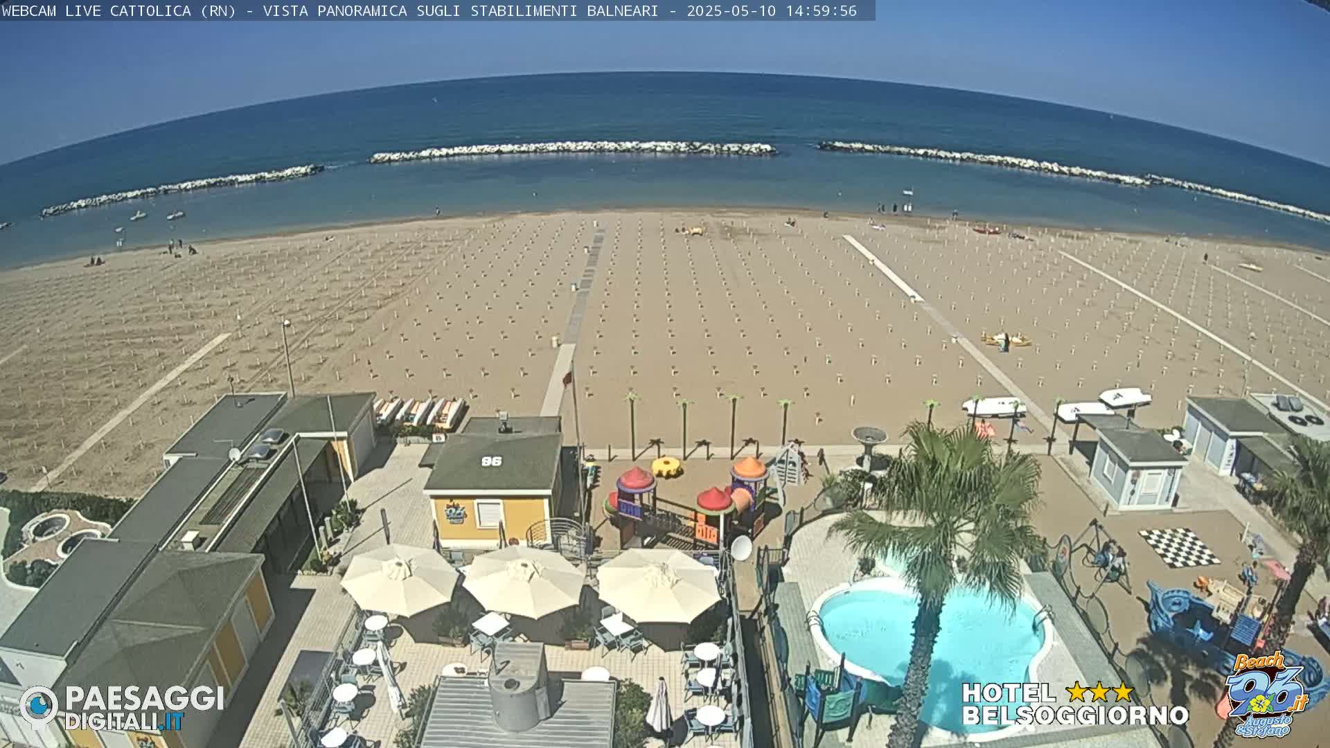 A mostly sunny day at a beach resort shows a sandy beach with rows of umbrellas and beach chairs, a swimming pool, and a playground near beachside buildings.